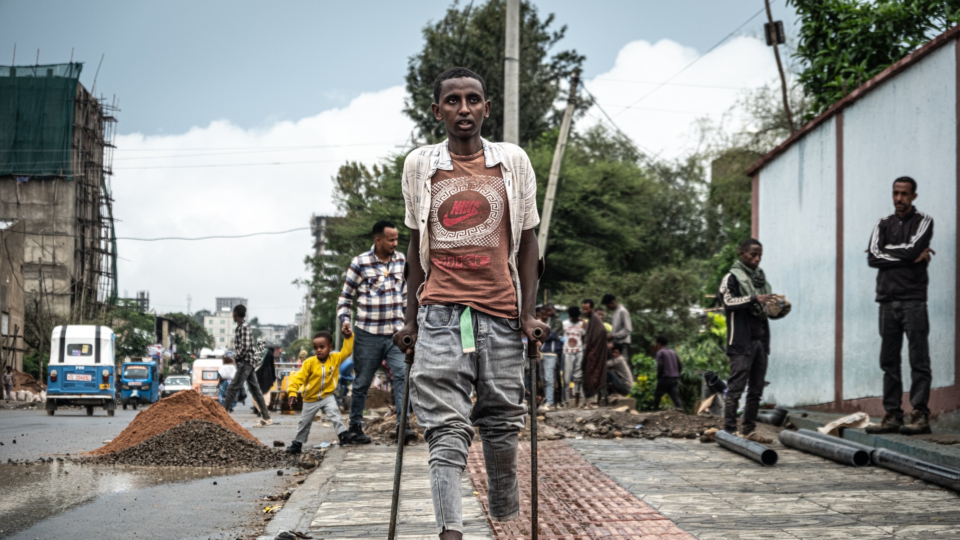 L'image montre une scène urbaine animée, où plusieurs personnes sont visibles dans un environnement de construction. Au premier plan, un jeune homme marche avec des béquilles, portant un t-shirt et un short. Il semble concentré et déterminé. En arrière-plan, on aperçoit d'autres travailleurs s'affairant dans la rue, entourés de matériaux de construction comme du gravier et des tuyaux. Le ciel est nuageux, suggérant une atmosphère légèrement grise. À droite, un mur peint est visible, ajoutant une touche de couleur au paysage de chantier.