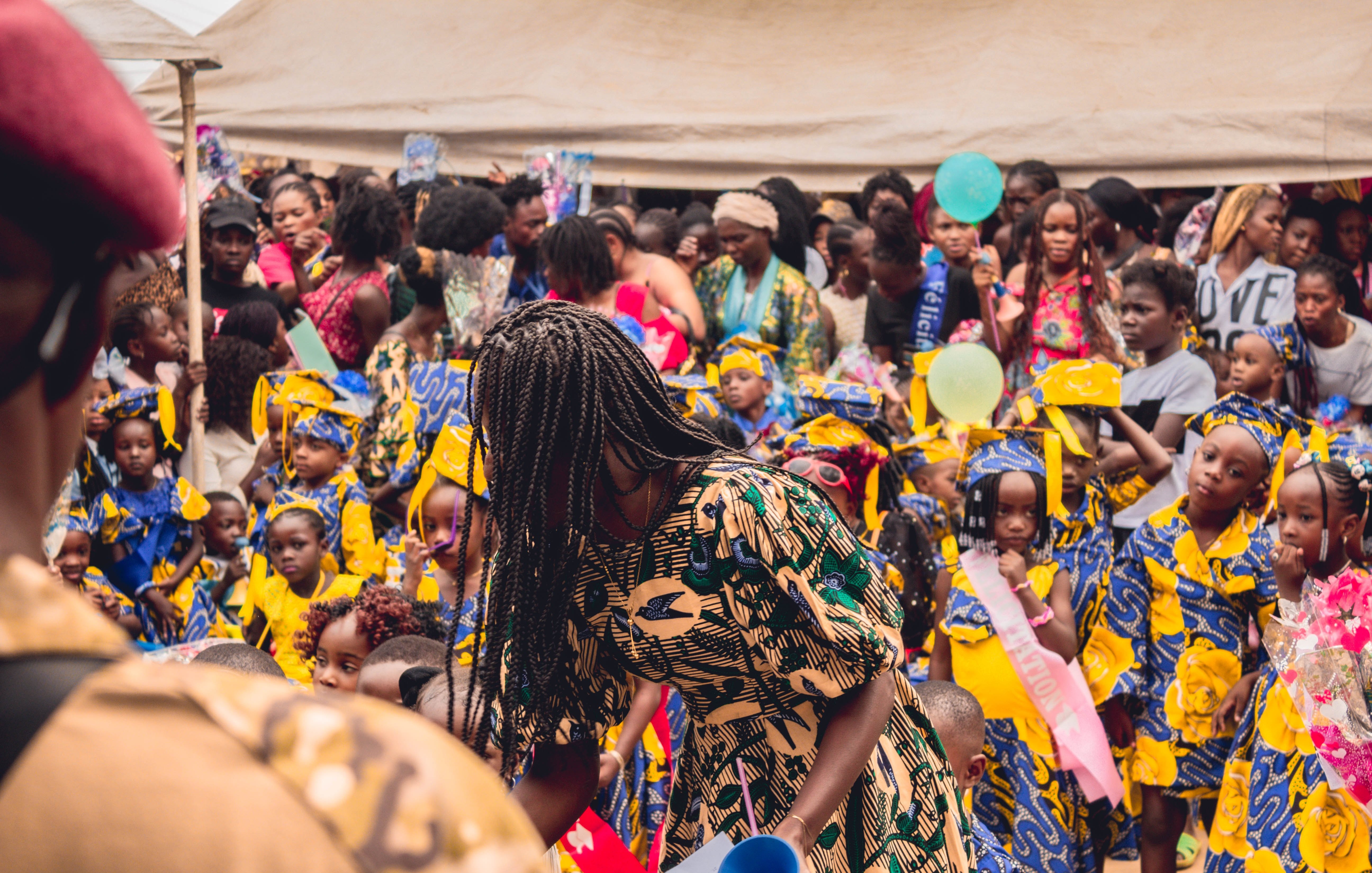 L'image montre une scène vibrante et animée d'un événement en plein air, probablement une célébration ou un festival. Au premier plan, on voit une femme portant une robe aux motifs colorés qui se penche, en train de distribuer quelque chose. Derrière elle, un grand groupe de filles, habillées de robes jaunes ornées de motifs bleus, se tient avec des sourires sur le visage. Leur ambiance semble joyeuse et festive. La foule est dense, avec de nombreuses personnes, y compris des femmes et des enfants, qui interagissent et profitent de l'événement. Des éléments décoratifs, comme des ballons ou des banderoles, pourraient ajouter une touche festive à l'environnement. L'atmosphère globale dégage une sensation de communauté et de célébration.
