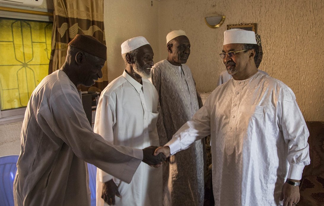 L'image montre un groupe de quatre hommes dans une pièce intérieure. Ils portent des vêtements traditionnels : des turbans et des vêtements clairs, souvent en coton. Un des hommes, sur la droite, est en train de serrer la main d'un autre homme, tandis que deux autres les regardent avec un air amical. La lumière entre par une fenêtre derrière eux, créant une atmosphère chaleureuse. Les murs sont texturés, avec un éclairage doux, et l'ensemble donne une impression de convivialité et de respect mutuel.