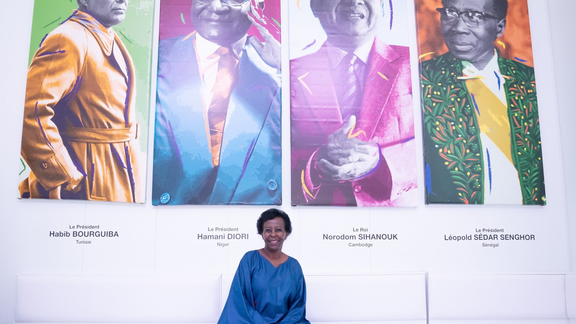 L'image présente une femme assise sur un banc dans une galerie moderne. Derrière elle, il y a quatre portraits colorés de figures politiques africaines. Chaque portrait est visiblement distinct, avec des couleurs vives, représentant des hommes portant des costumes traditionnels ou modernes. La femme, vêtue d'une robe bleu foncé, sourit légèrement et semble apprécier l'exposition. Le décor est minimaliste, avec des sièges blancs et des poufs de différentes couleurs qui ajoutent une touche de dynamisme à l'ensemble. L'ambiance générale est accueillante et artistique, invitant à la réflexion sur l'histoire politique de l'Afrique.