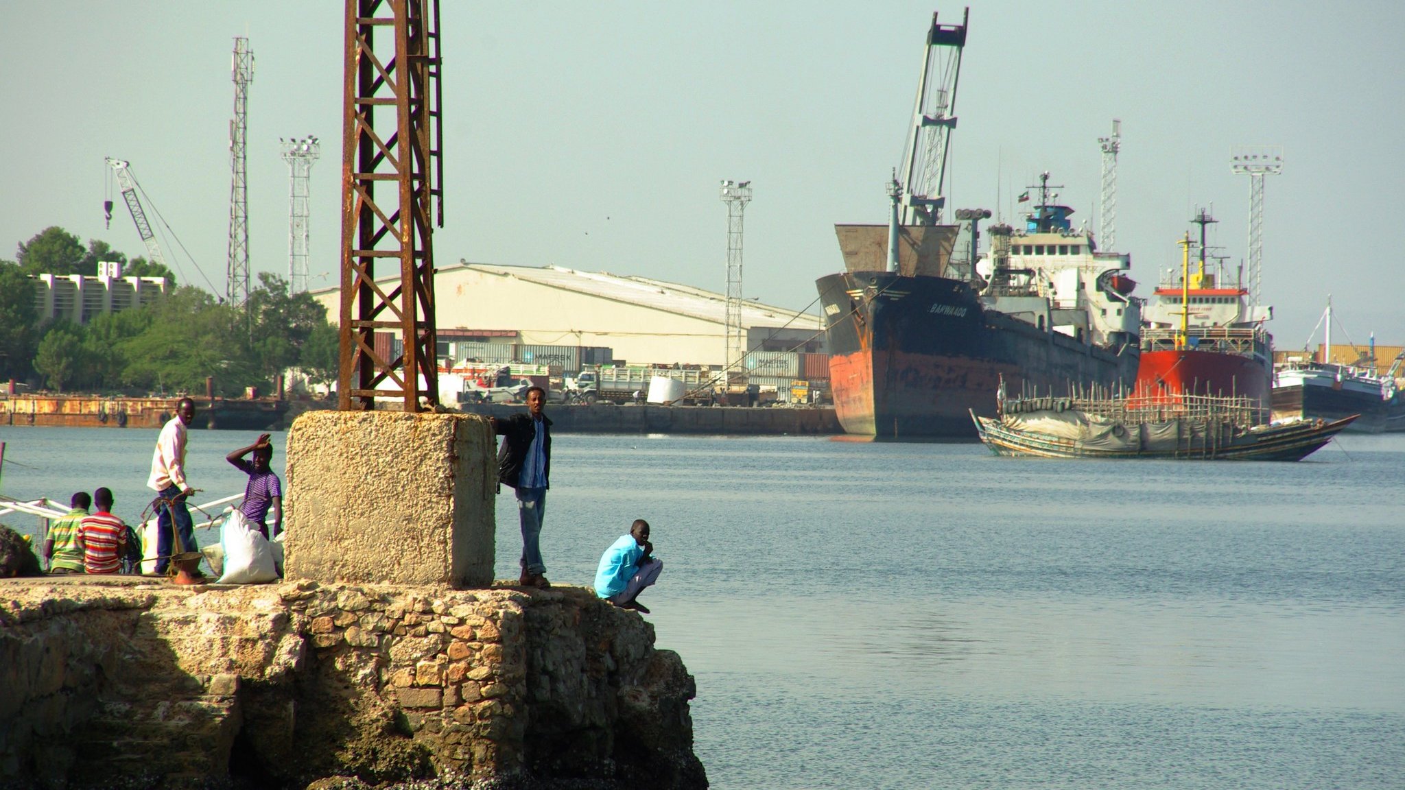 Imaginez un port animé. Au premier plan, il y a un quai en pierre avec plusieurs personnes : certaines sont debout, discutant, tandis que d'autres se penchent sur le bord pour observer l'eau. L'atmosphère est vivante, remplie d'échos de rires et de conversations. À l'arrière-plan, de grands navires sont amarrés, leurs coques visibles au-dessus de l'eau paisible. Des grues et des poteaux élancés se dressent, témoignant des activités portuaires. L'air sent légèrement le sel et le poisson, créant une ambiance maritime authentique.
