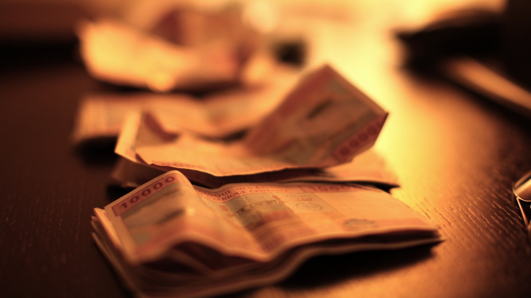 L'image montre un plan rapproché de billets de banque étalés sur une surface sombre, probablement une table en bois. Les billets sont flous, suggérant une légère profondeur de champ, ce qui attire l'attention sur la texture et les détails des billets. L'éclairage est doux et chaud, avec une lumière diffuse provenant d'une source hors du champ de vision, créant une ambiance intime et feutrée. Cette lumière fait ressortir les motifs et les couleurs des billets, ajoutant une dimension visuelle à l'ensemble.