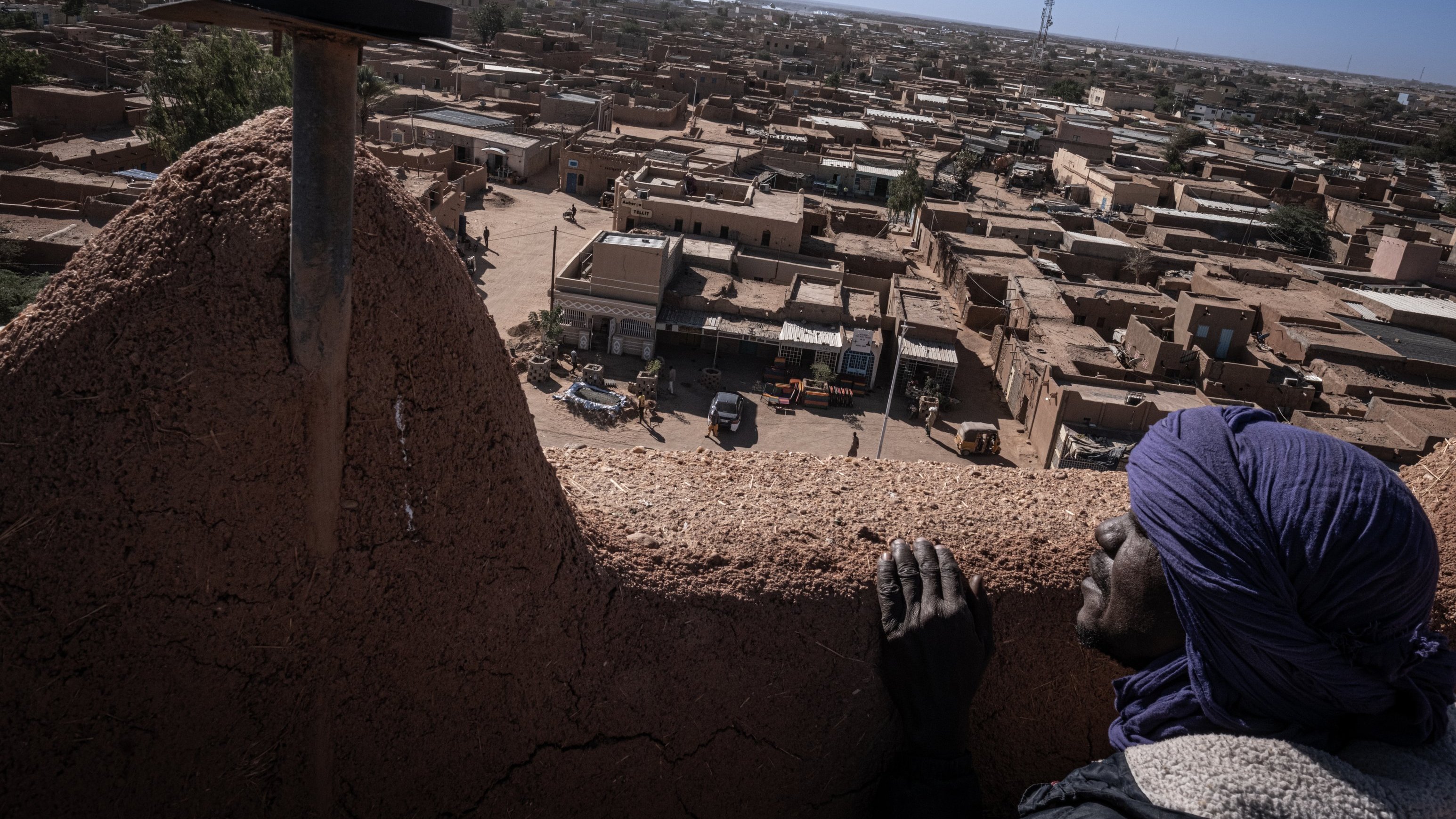 L'image présente une vue panoramique d'une ville saharienne. Au premier plan, une personne au visage marqué par le temps s'appuie sur un mur en terre. Elle porte un turban et semble contempler le paysage. En arrière-plan, des maisons en adobe s'étendent à perte de vue, avec des toits plats et des teintes ocres. On peut apercevoir des petites routes et des véhicules qui circulent. L'horizon est dégagé, avec quelques structures modernes qui contrastent avec l'architecture traditionnelle. L'atmosphère évoque à la fois la chaleur du désert et la tranquillité d'une ville endormie.
