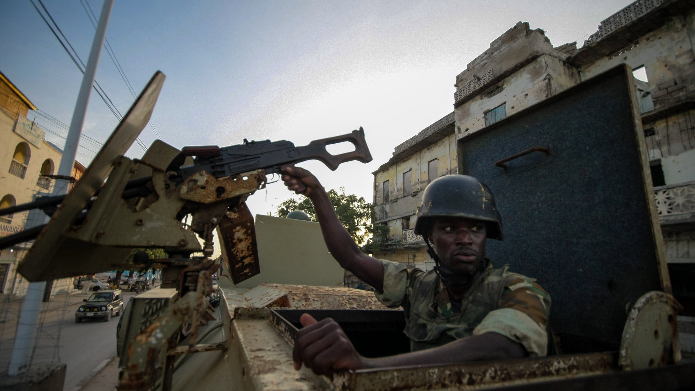 L'image montre un soldat assis dans un véhicule militaire. Il est vêtu d'un uniforme camouflage et porte un casque. Le soldat tient un fusil d'assaut, que l'on peut imaginer prêt à être utilisé. En arrière-plan, on aperçoit des bâtiments en ruine, suggérant que l'environnement est marqué par des signes de conflit. La lumière est douce, indiquant peut-être un lever ou un coucher de soleil. L'atmosphère dégage un sentiment de tension et de vigilance dans un cadre urbain dégradé.