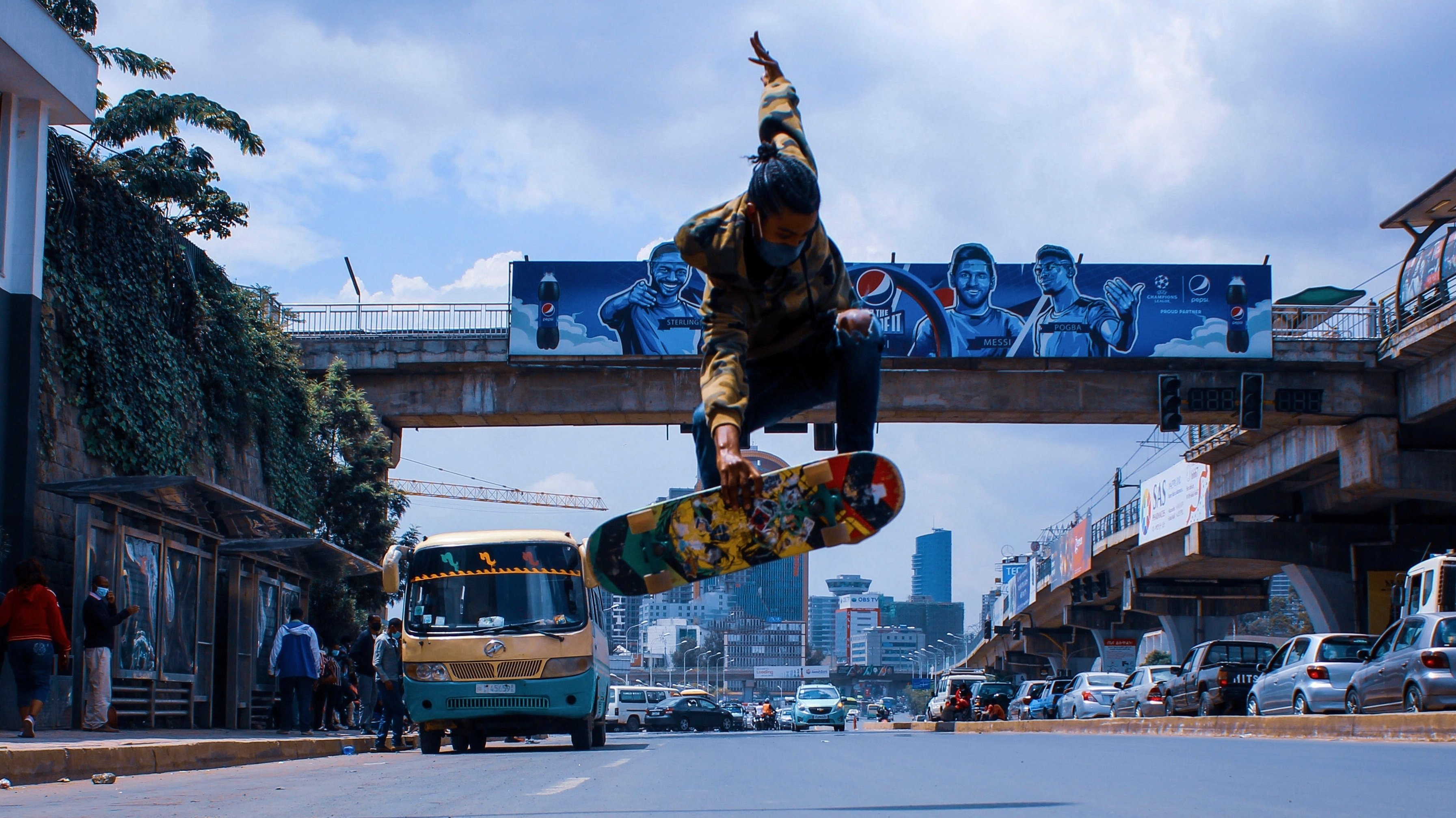 L'image montre un skateur en plein saut au-dessus de la route. Il est habillé d'un sweat à capuche avec des motifs et porte un skateboard coloré. En arrière-plan, il y a des bâtiments urbains, des graffitis sur un mur, et un bus qui passe, typique des grandes villes. La scène capture une ambiance dynamique et énergique, avec un ciel partiellement nuageux qui ajoute à l'atmosphère urbaine. Les paysages urbains et la présence d'autres personnes montrent l'animation de la ville.