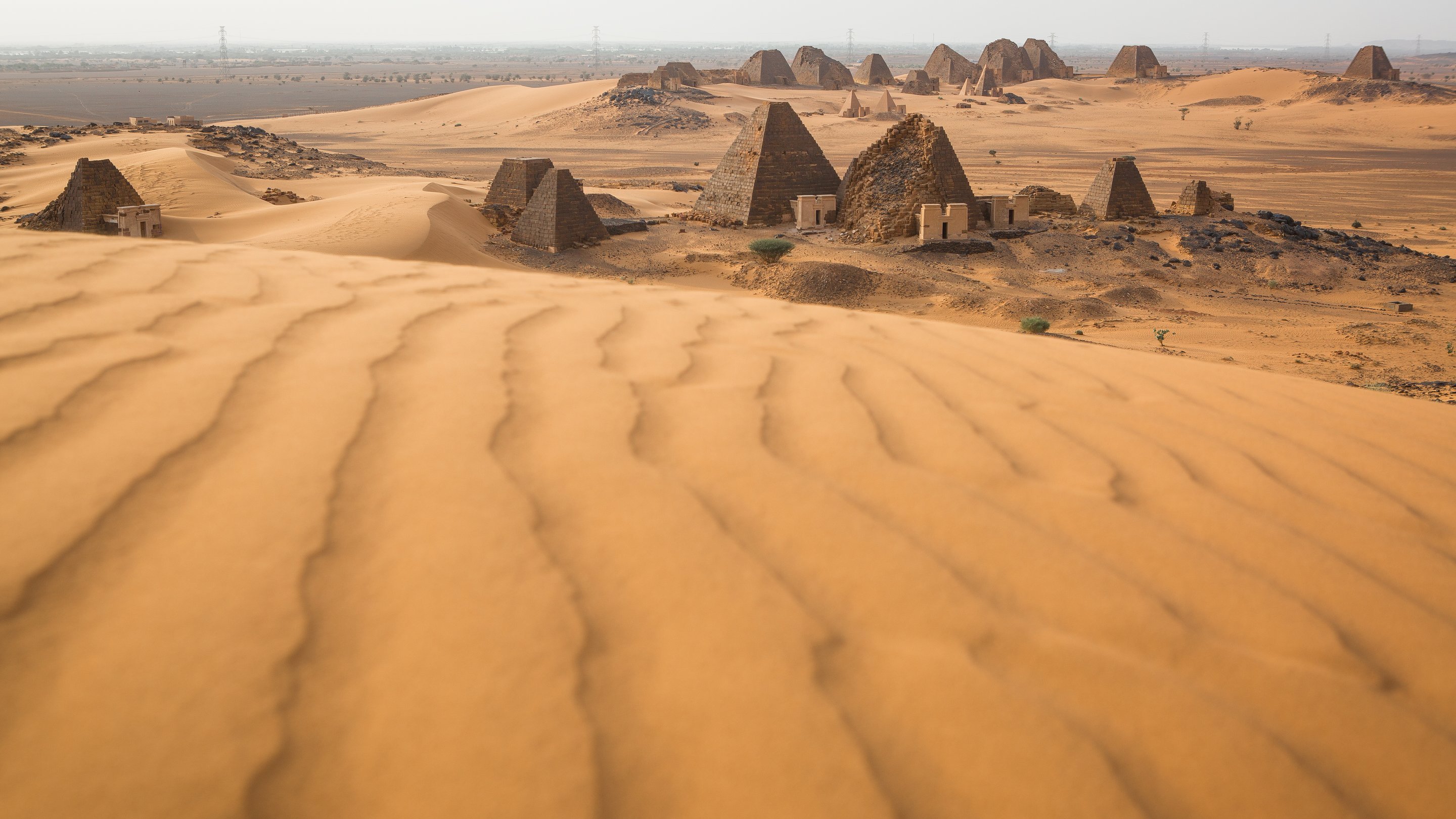 L'image décrit un paysage désertique, dominé par de vastes dunes de sable aux formes ondulantes. En arrière-plan, on aperçoit des pyramides anciennes dont les sommets sont pointus et les surfaces sont qui semblent rugueuses. Le terrain autour des pyramides est également parsemé de petites éboulis et de dunes, créant un paysage à la fois majestueux et mystérieux. La lumière du soleil éclaire la scène, mettant en valeur les nuances dorées du sable et l'architecture historique des pyramides. L'atmosphère générale évoque la chaleur et l'immensité du désert, ainsi qu'un sentiment d'émerveillement face aux vestiges du passé.