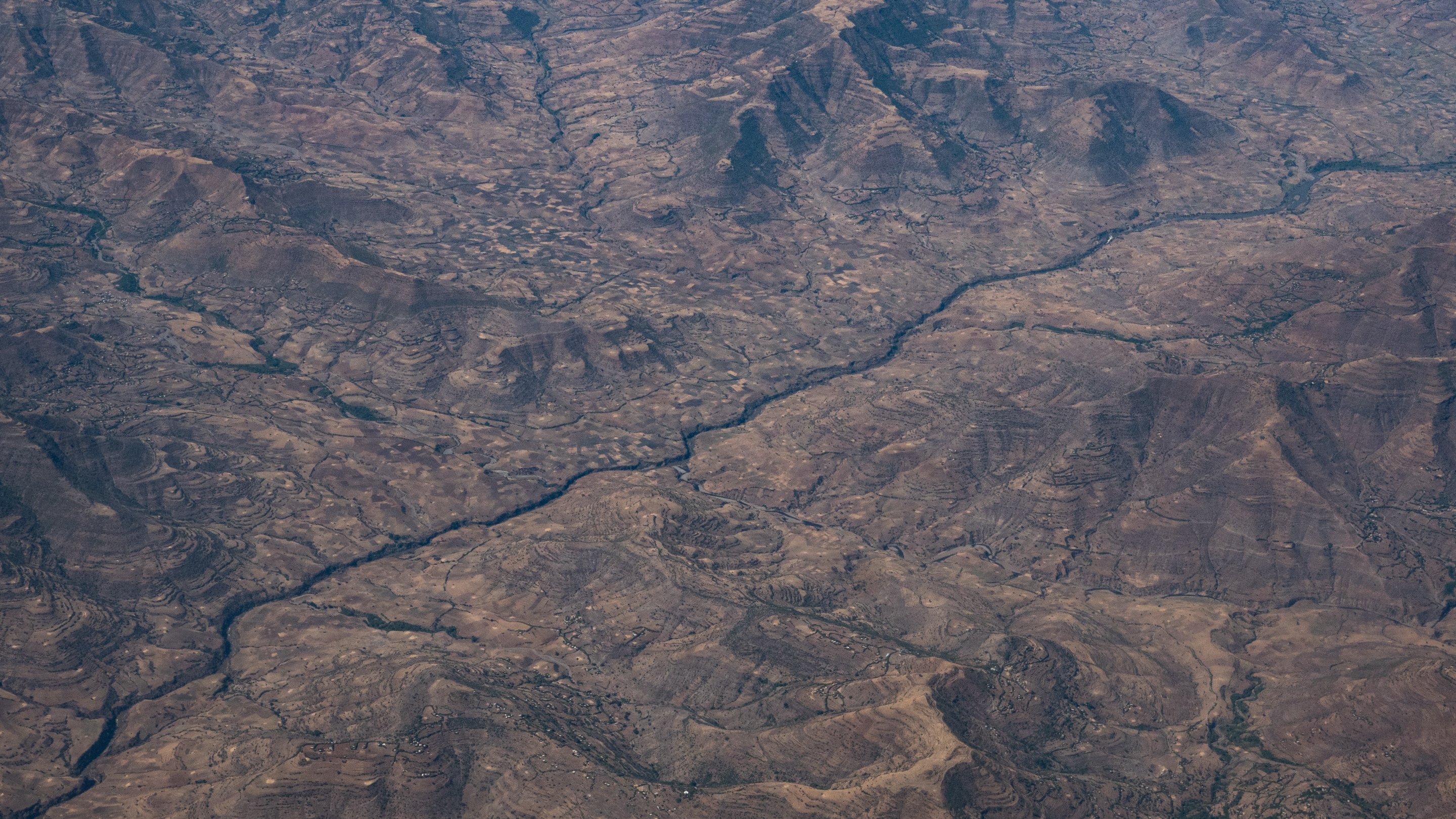 Cette image montre un paysage désertique vu du ciel. On peut observer de vastes étendues de terrain aride, striées par des vallées et des collines aux formes irrégulières. Au centre, un cours d'eau serpente à travers ce paysage, créant un contraste avec les zones sèches environnantes. Les couleurs dominantes sont des teintes de marron et de beige, évoquant un sol rocailleux et dénudé, tandis que la rivière apporte une touche de reflets plus sombres, suggérant des zones humides dans ce milieu. L'ensemble dégage une impression de vastitude et de sérénité, avec une nature sauvage encore peu altérée par l'homme.