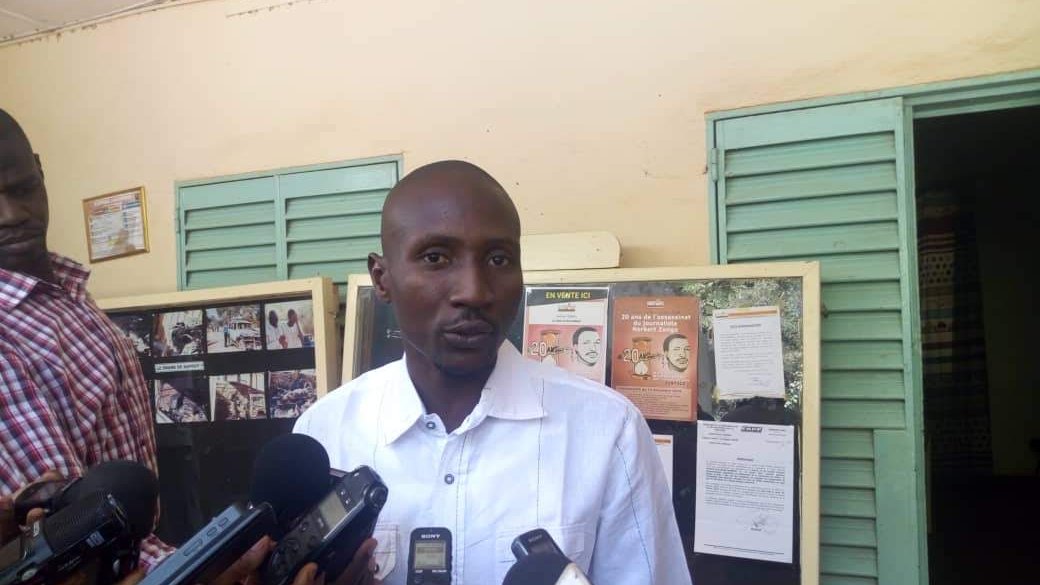 Dans cette image, un homme se tient devant un mur avec des panneaux d'affichage. Il porte une chemise blanche et semble parler à plusieurs journalistes qui l'entourent, tenant des microphones et des appareils d'enregistrement. Les panneaux d'affichage derrière lui contiennent des informations, des photos et des affiches. L'ambiance paraît être celle d'une conférence de presse ou d'un événement public où des informations sont partagées.