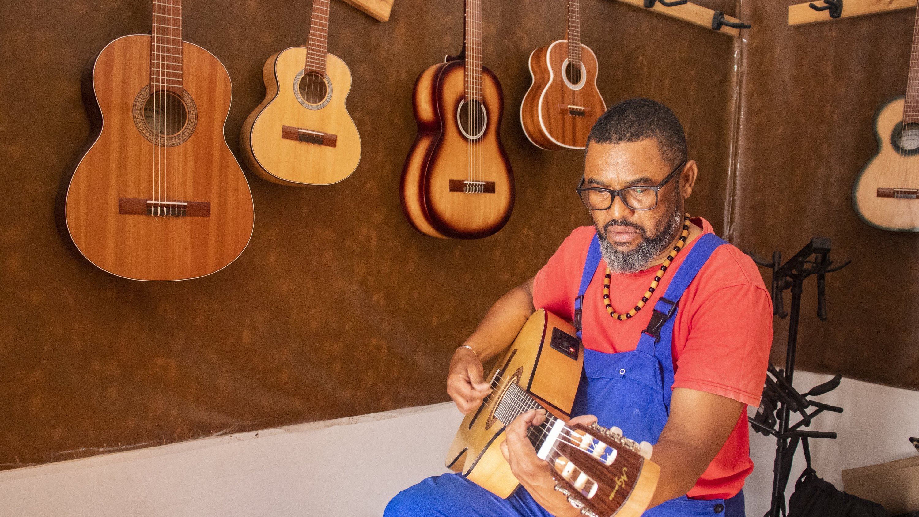 L'image montre un homme assis, vêtu d'une salopette bleue et d'un t-shirt rouge. Il est en train de jouer de la guitare acoustique avec une expression de concentration sur son visage. Autour de lui, plusieurs guitares sont accrochées au mur, dans des teintes de bois naturel. L'ambiance de l'espace est chaleureuse et artistique, soulignant sa passion pour la musique.