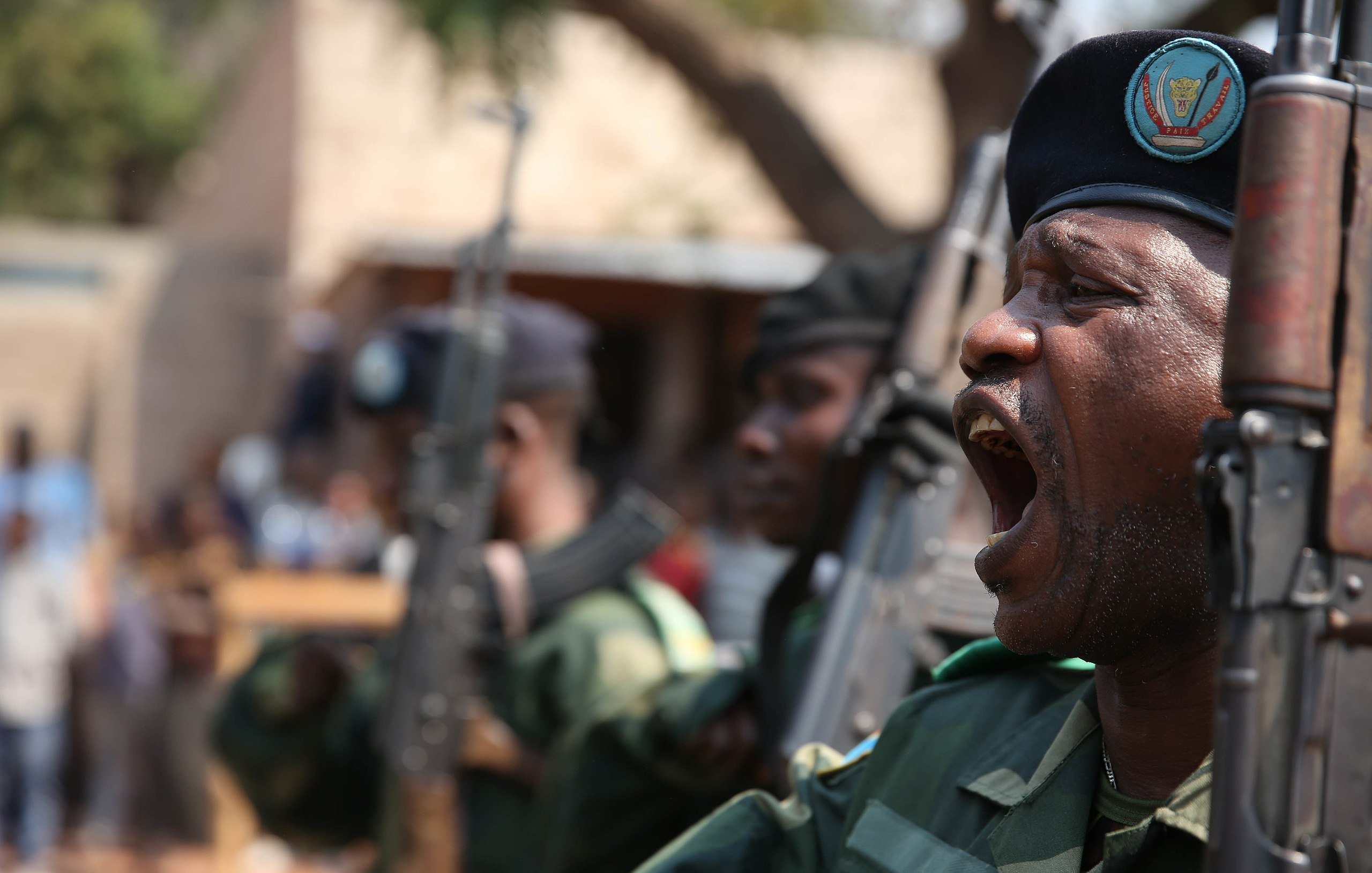 L'image montre un groupe de militaires en uniforme, en position de parade. L'un d'eux, au premier plan, est en train de crier ou de donner un commandement, affichant une expression de détermination. Ils portent des bérets sombres et tiennent des fusils en position verticale. En arrière-plan, on peut apercevoir d'autres soldats ainsi qu'un groupe de personnes qui semblent regarder la scène. L'environnement est extérieur, probablement lors d'un événement officiel ou d'une cérémonie. L'atmosphère est solennelle et dynamique, évoquant un sentiment de force et de discipline.