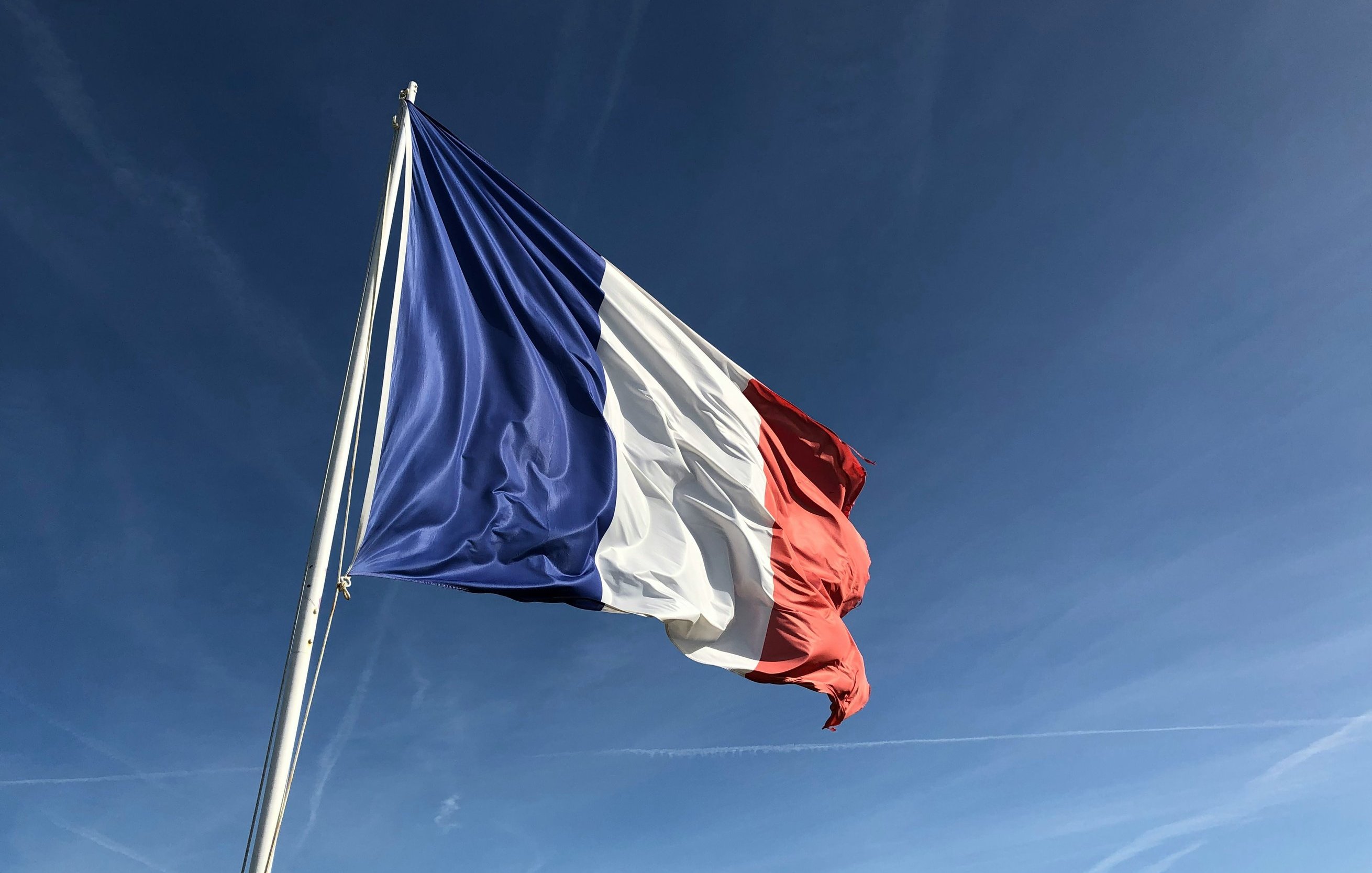 L'image montre un drapeau, qui est le drapeau tricolore français. Il est composé de trois bandes verticales : bleu à gauche, blanc au centre et rouge à droite. Le drapeau est agité par le vent, ce qui donne une impression de mouvement. À l'arrière-plan, on peut imaginer un ciel clair, probablement d'une belle journée ensoleillée, avec quelques traînées de nuages dans un bleu vif. Le contraste entre les couleurs du drapeau et la luminosité du ciel crée une image frappante et symbolique.