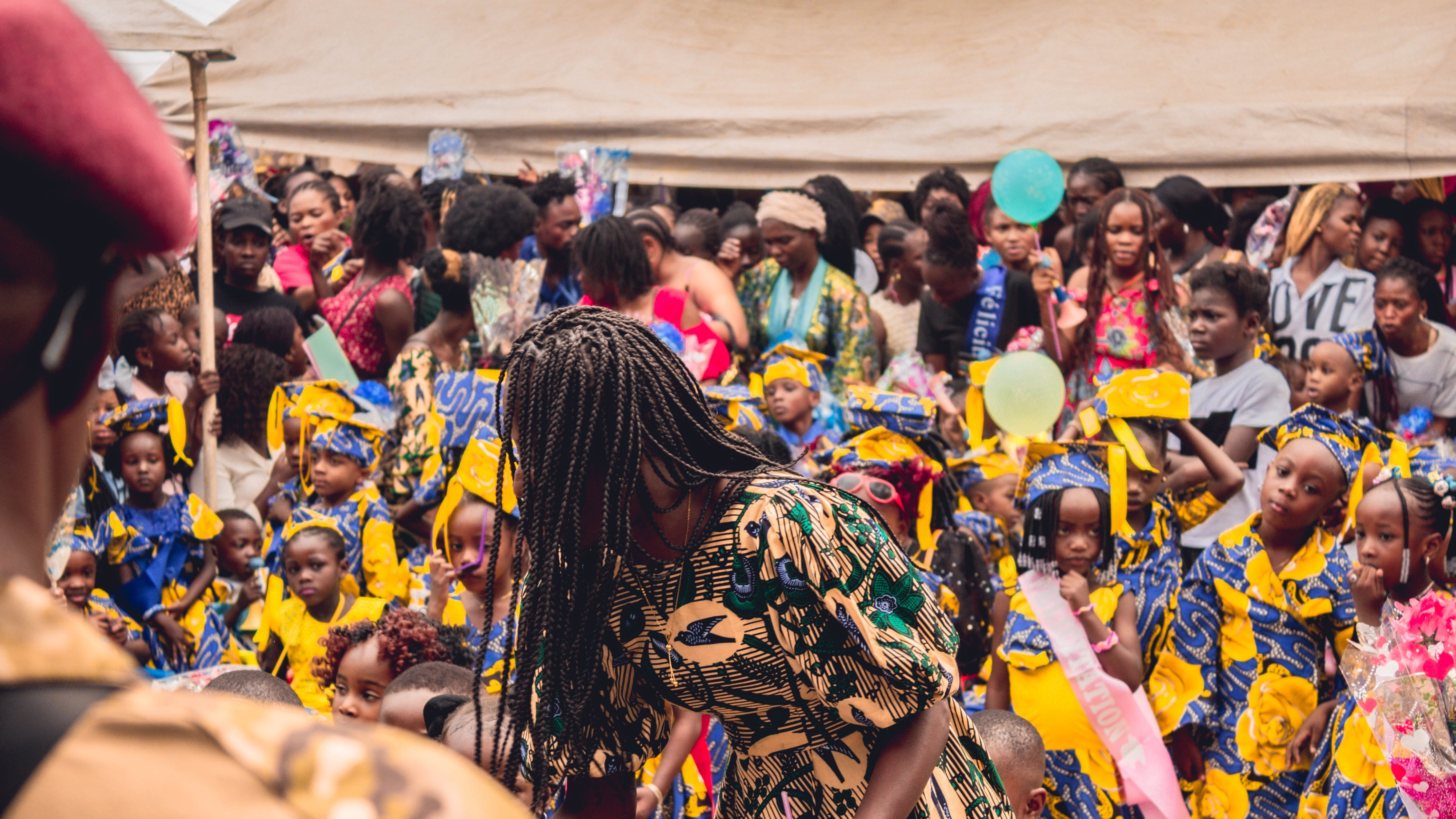 L'image montre une scène vibrante et animée d'un événement en plein air, probablement une célébration ou un festival. Au premier plan, on voit une femme portant une robe aux motifs colorés qui se penche, en train de distribuer quelque chose. Derrière elle, un grand groupe de filles, habillées de robes jaunes ornées de motifs bleus, se tient avec des sourires sur le visage. Leur ambiance semble joyeuse et festive. La foule est dense, avec de nombreuses personnes, y compris des femmes et des enfants, qui interagissent et profitent de l'événement. Des éléments décoratifs, comme des ballons ou des banderoles, pourraient ajouter une touche festive à l'environnement. L'atmosphère globale dégage une sensation de communauté et de célébration.