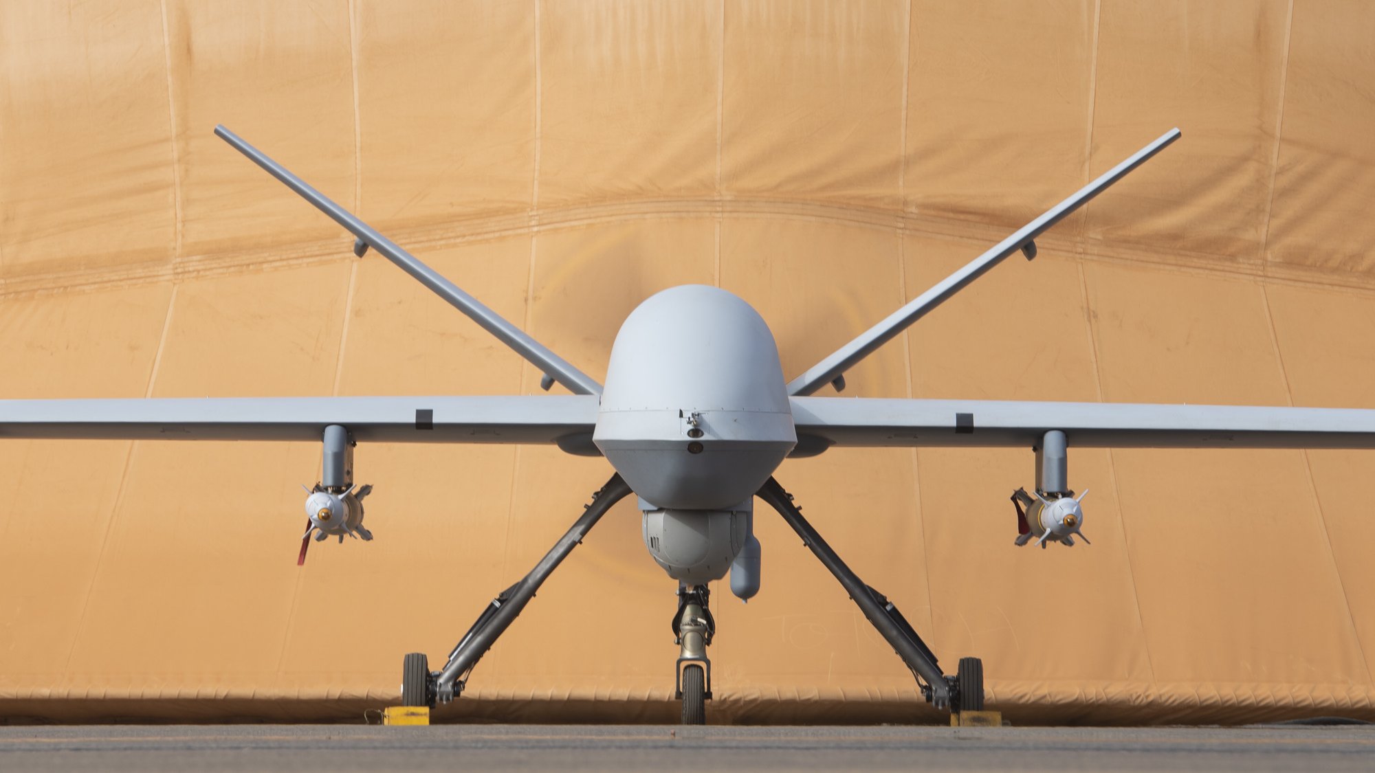 L'image montre un drone militaire de type MQ-9 Reaper. Il est positionné de face, avec le corps fuselé et l'aile légèrement étendue. À chaque côté du fuselage, on peut voir des réservoirs ou des munitions. En arrière-plan, il y a une toile beige qui semble un hangar ou une protection, lui donnant un aspect de préparation à un vol. Le sol est en béton, créant un environnement d'aérodrome. L'ensemble est impressionnant, évoquant la technologie aéronautique moderne.