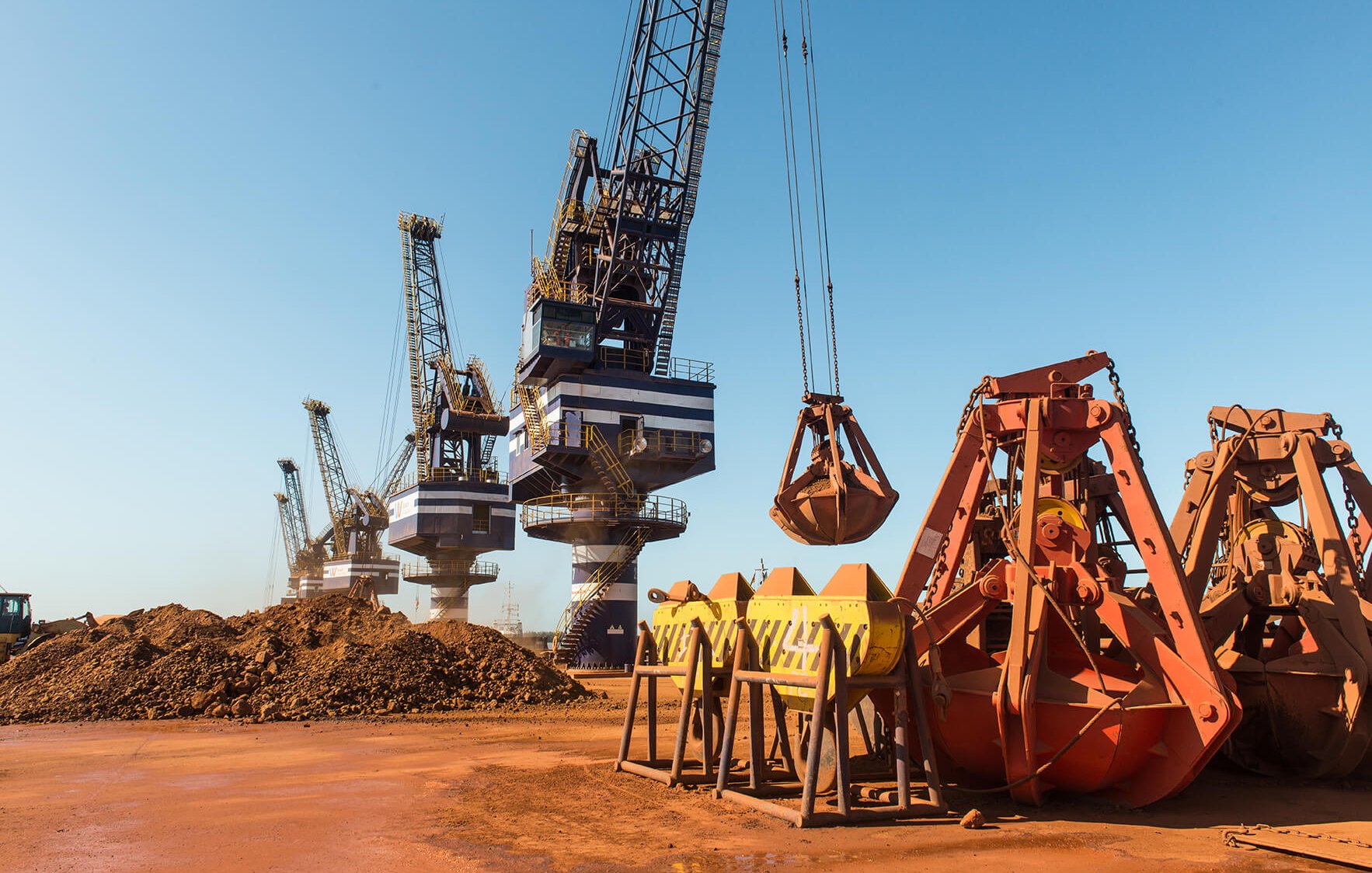 Dans cette image, nous sommes dans un port industriel où plusieurs grues sont visibles. Ces grandes machines sont utilisées pour charger et décharger des matériaux. À l'avant, il y a des godets et des bennes orange, qui sont des équipements de levage, reposant sur le sol. En arrière-plan, plusieurs grues se dressent contre un ciel bleu clair, leurs bras et leurs crochets descendus vers les tas de terre brune. L'atmosphère est typiquement industrielle, avec un sol en terre battue et une ambiance de travail intense. Des sons de machines en fonctionnement et le bruit des cargaisons en mouvement pourraient être présents dans cet environnement.