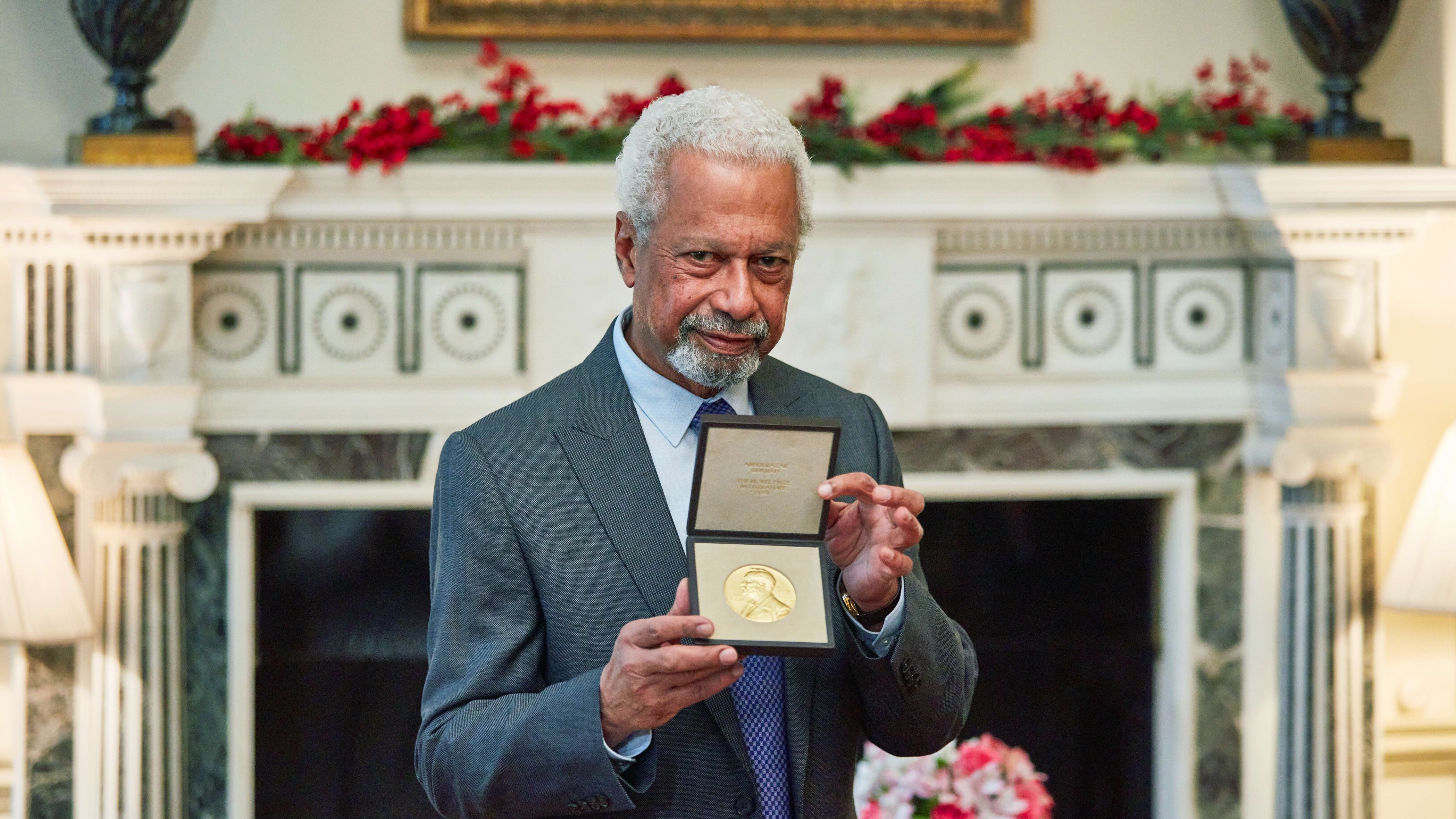 L'image montre un homme âgé, avec des cheveux poivre et sel et une barbe, qui porte un costume gris. Il se tient fièrement devant une cheminée décorée, tenant une médaille en or dans un écrin, qu'il montre à la caméra. En arrière-plan, on peut distinguer une décoration florale avec des fleurs disposées sur une table, créant une ambiance chaleureuse et élégante. La pièce est bien éclairée et a un air prestigieux, suggérant une occasion spéciale.
