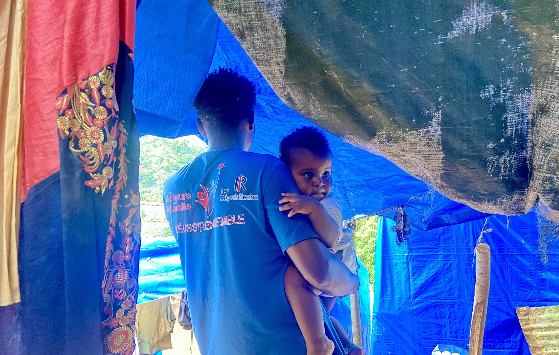 L'image montre un homme portant un enfant dans ses bras. Ils se trouvent sous une tente improvisée faite de tissus aux couleurs vives comme le bleu et le rouge. Le décor semble rural, avec des éléments naturels en arrière-plan. L'homme a un t-shirt bleu avec des inscriptions, et l'enfant, qui regarde légèrement en arrière, semble être de très jeune âge. La scène évoque un moment de tendresse et de protection dans un environnement simple.