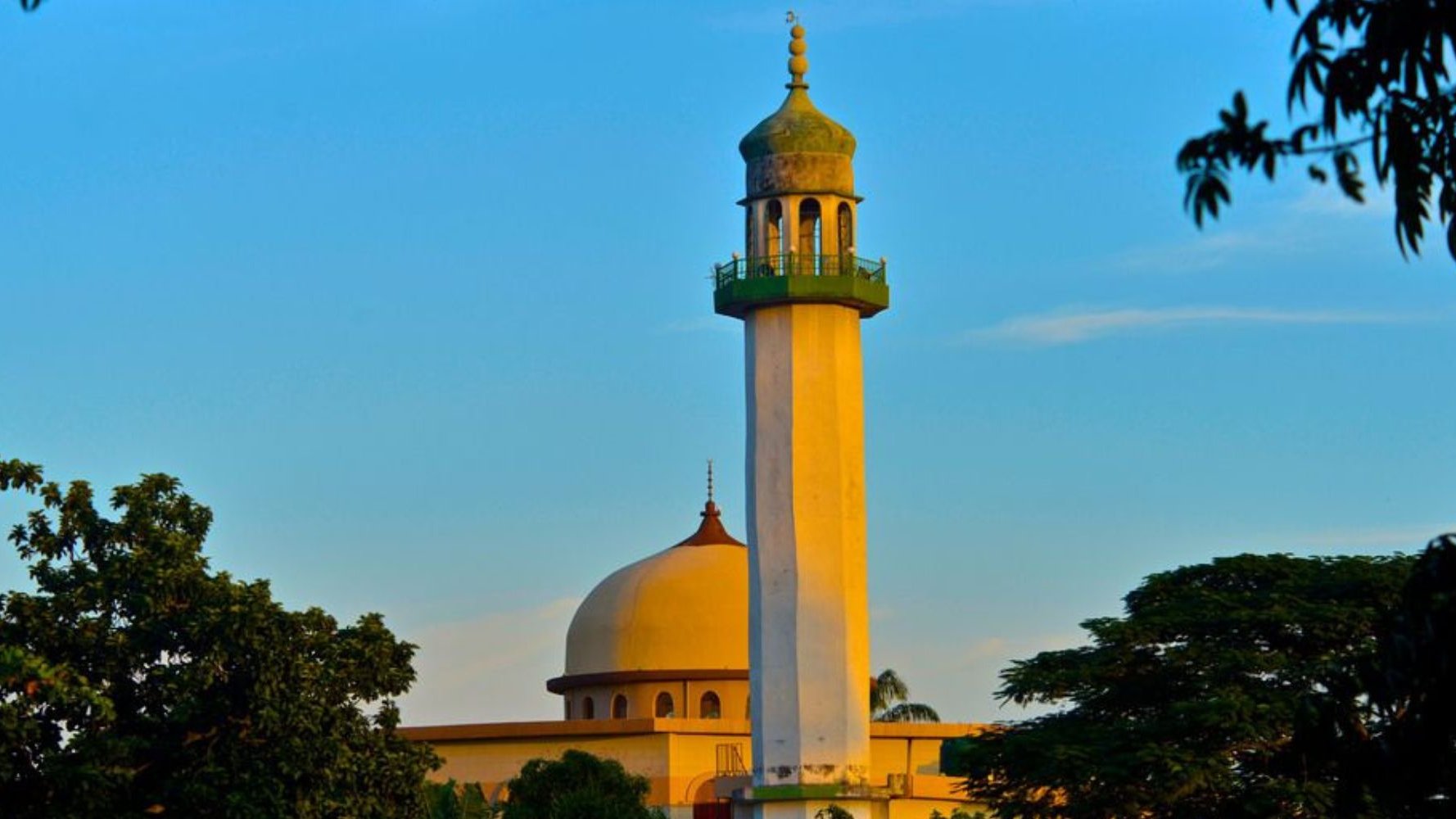 L'image montre un groupe de personnes en train de prier ensemble. Au centre, un homme en costume bleu se tient les mains jointes, la tête légèrement inclinée, montrant une attitude de recueillement. Autour de lui, plusieurs hommes portent des vêtements traditionnels, incluant des turbans et des chapeaux décorés. Tous semblent concentrés et tournés vers l'avant, les yeux fermés ou la tête baissée, dans un moment de méditation ou de prière. L'ambiance est empreinte de sérénité et de respect.
