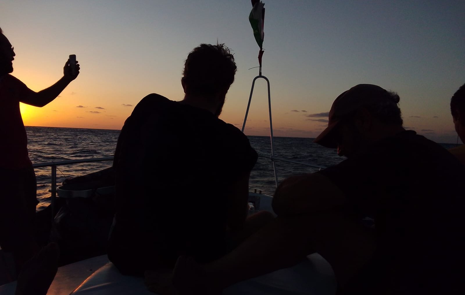 Imaginez une scène sereine sur l'eau, à l'heure où le soleil se couche. Des silhouettes de personnes se découpent contre un fond doré. À gauche, une personne tient un téléphone, capturant le moment, tandis que d'autres sont assises, tournées vers l'horizon. Leurs formes sont sombres, presque spectrales, créant un contraste avec les couleurs chaudes du ciel qui passent du jaune à l'orange et au mauve. La surface de l'eau scintille doucement, reflétant les teintes du crépuscule. L'atmosphère est calme, empreinte de contemplation et d'amitié, tandis que le paysage maritime s'étend à perte de vue.