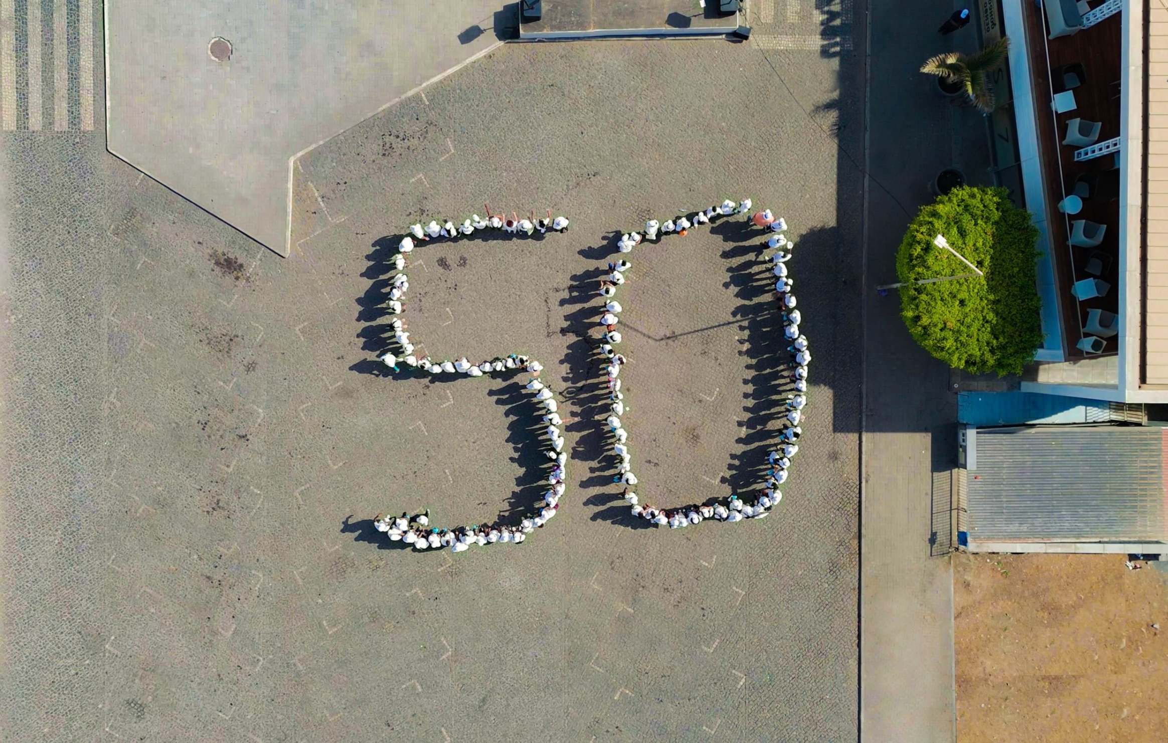 L'image montre une vue aérienne d'une place où des personnes sont réunies pour former le chiffre "50". Les participants sont disposés de manière à créer les deux chiffres avec leurs corps, et ils semblent porter des vêtements clairs, peut-être des t-shirts blancs. Autour, on peut voir des pavés au sol et quelques éléments de paysage urbain, comme des arbres et des bâtiments en arrière-plan. L'ambiance semble conviviale et festive, suggérant que cet événement marque une célébration ou un anniversaire.