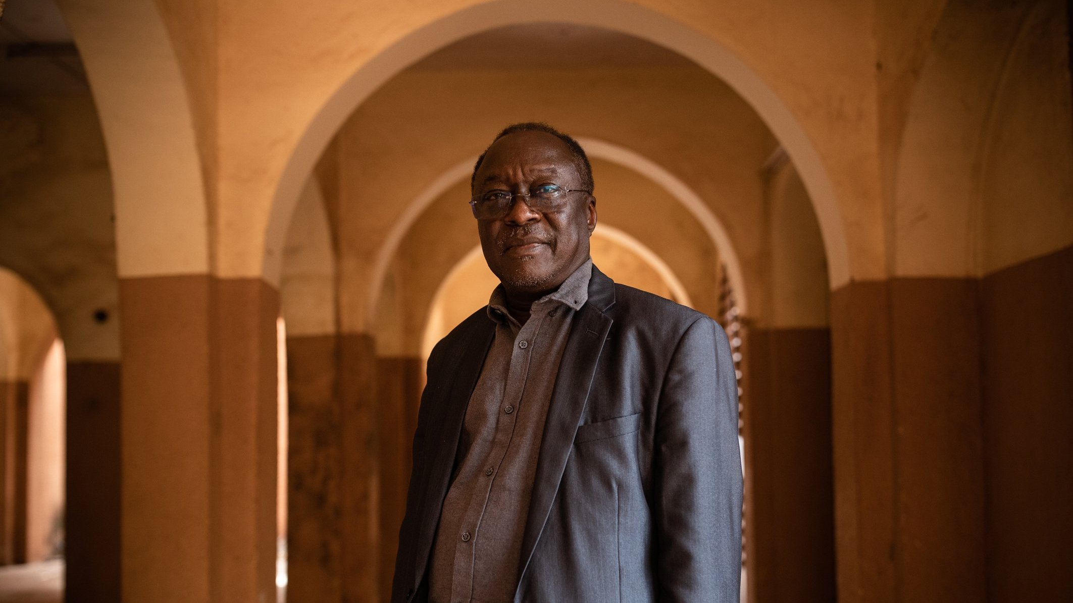 L'image montre un homme debout dans un espace intérieur, caractérisé par de grandes arcades. Les murs sont peints de couleurs chaudes, avec des tons ocre et beige. L'homme porte une veste sombre et une chemise grise, et il a l'air confiant. Son expression est sereine et il regarde légèrement vers la droite. L'arrière-plan présente des arches qui créent une atmosphère de profondeur et de perspective. La lumière semble douce et diffuse, ajoutant une ambiance chaleureuse à la scène.