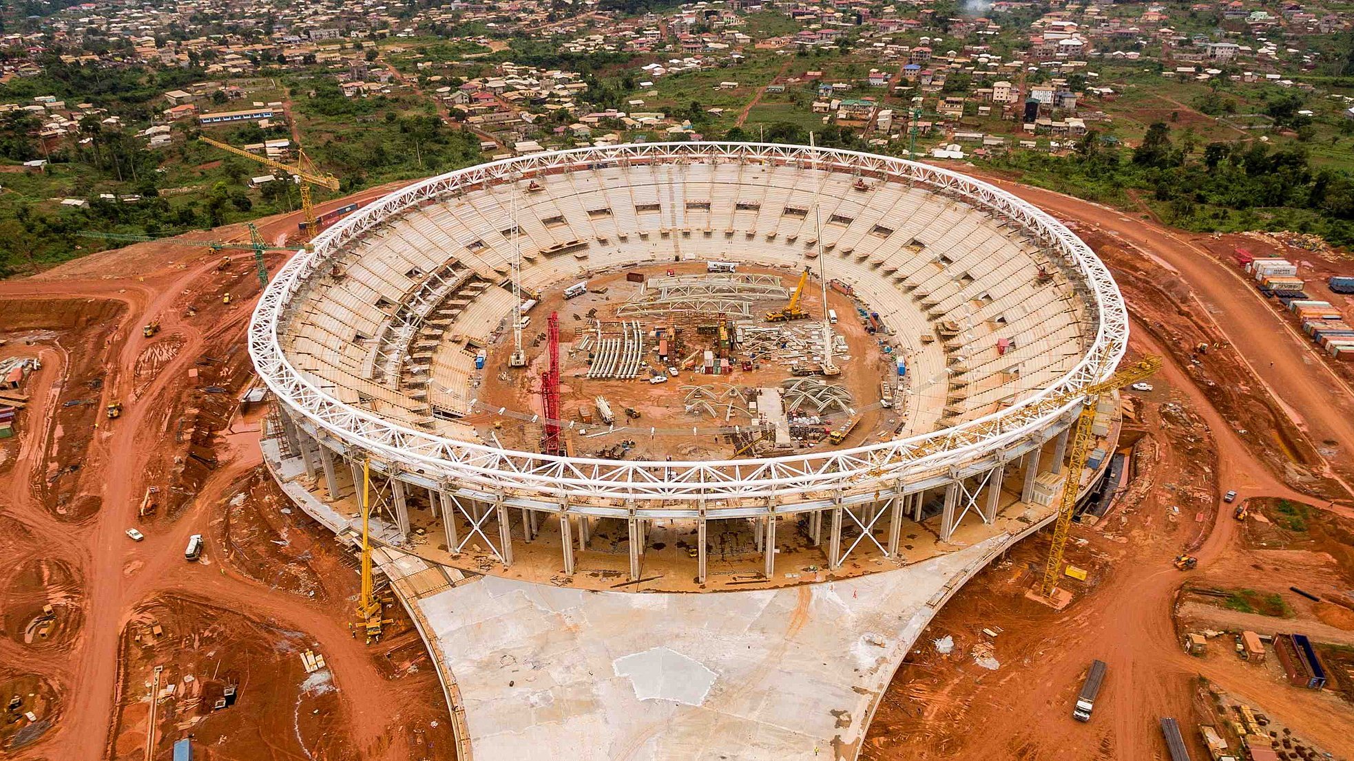 L'image montre un stade en cours de construction, vu d'en haut. Le stade a une forme ovale et un grand espace intérieur qui est encore vide, avec des structures en cours d'installation. Autour du stade, on peut voir un terrain rouge, probablement une terre fouillée pour la construction. À l'extérieur de la structure, il y a des machines de construction et des matériaux éparpillés. À l'arrière-plan, des maisons et des collines sont visibles, ce qui donne une idée de l'environnement urbain et naturel qui entoure le stade. L'ensemble de la scène évoque une ambition de développement et de modernisation.