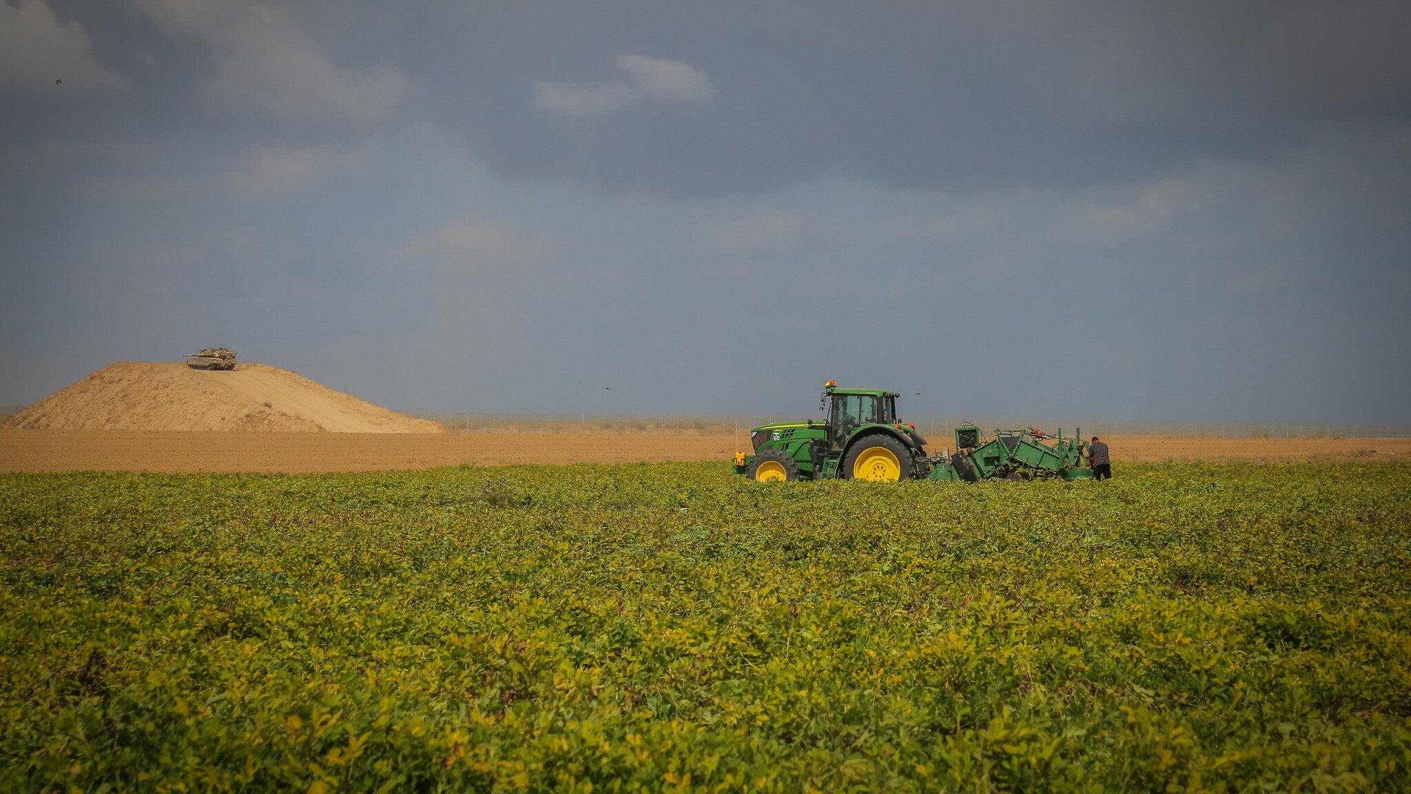 L'image présente un vaste champ de culture où l'on peut voir un tracteur vert, typique des fermes. Le tracteur est engagé dans une activité de labour, préparant le sol pour la plantation. En arrière-plan, on aperçoit une colline de terre argileuse, surmontée d'un véhicule. Le ciel est nuageux, créant une ambiance contrastée avec des teintes de bleu et de gris qui donnent une impression de calme. Les lignes horizontales des cultures et de la colline ajoutent une profondeur visuelle au paysage rural.