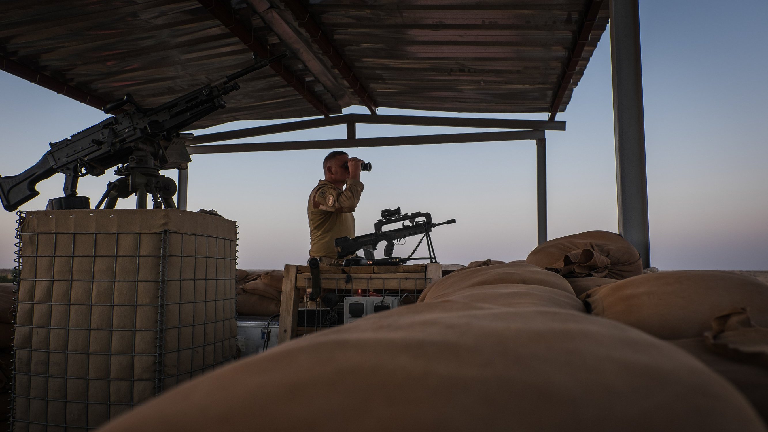 L'image montre un soldat en position d'observation sur un poste de garde en hauteur. Il se tient debout, avec des jumelles à la main, scrutant l'horizon. À sa droite, on aperçoit une mitrailleuse montée sur un support. Le sol est recouvert de sacs de sable, utilisés comme protection. L'éclairage de la scène évoque un moment de la journée où le ciel est teinté de couleurs douces, créant une atmosphère calme malgré le contexte militaire. Le soldat porte un uniforme et semble attentif à son environnement.