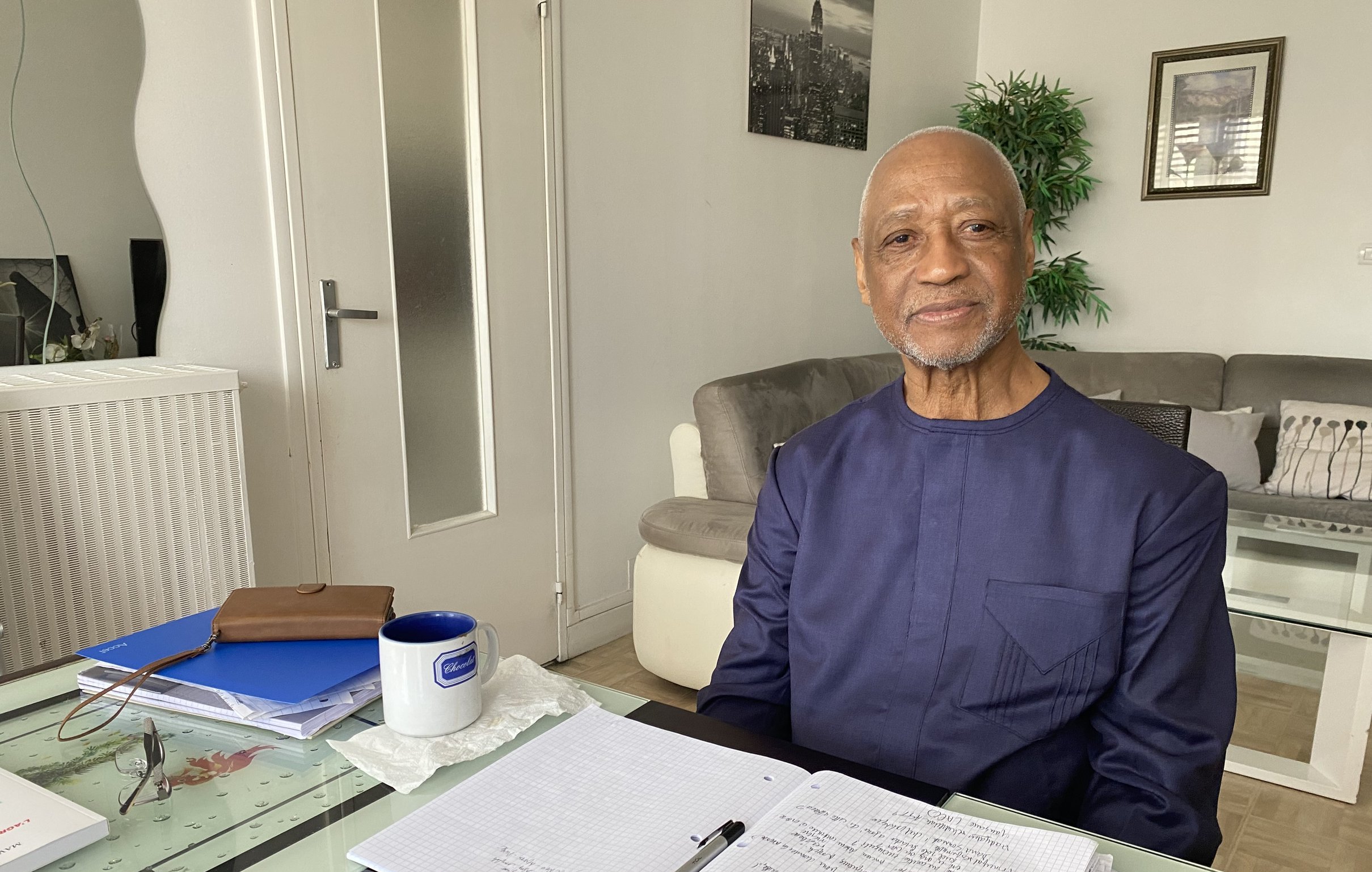L'image montre un homme assis à une table chez lui. Il porte une tenue traditionnelle de couleur sombre et affiche un sourire chaleureux. La table est en verre et est ornée de divers objets, dont un pot de fleurs vert, un carnet ouvert avec des notes, et un stylo. À côté du carnet, il y a une tasse bleue sur la table. En arrière-plan, on peut voir un canapé clair et des décorations murales. La lumière naturelle entre par une fenêtre, créant une ambiance lumineuse et accueillante.