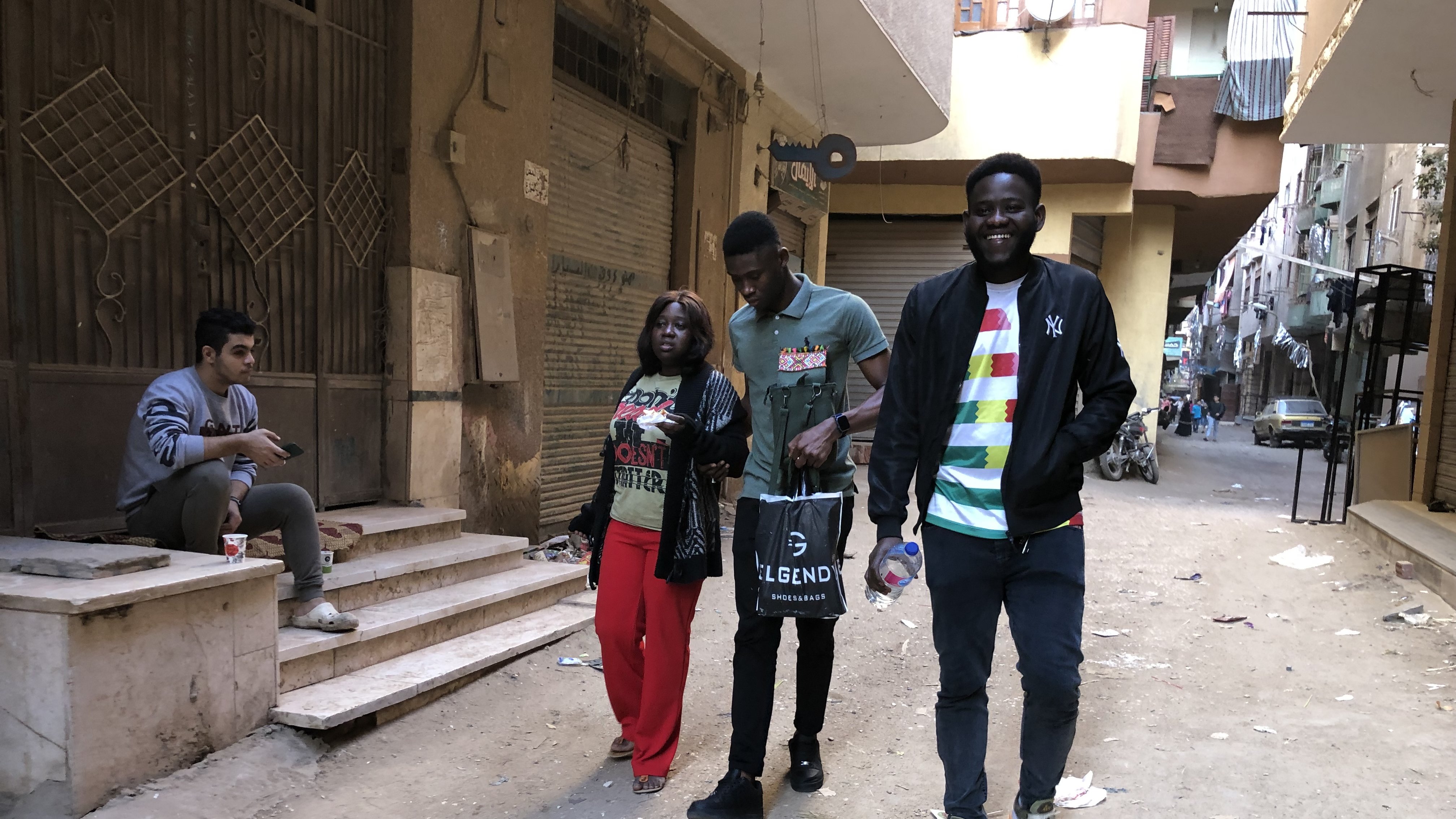 L'image montre une rue étroite et animée dans un quartier urbain. Il y a des déchets dispersés sur le sol, indiquant un environnement un peu désordonné. Sur le côté gauche, un jeune homme est assis sur des escaliers, absorbé par son téléphone ou ses réflexions. Au centre, trois personnes marchent ensemble : une femme portant un haut coloré et un pantalon rouge, un homme en t-shirt gris foncé et un autre homme vêtu d'un haut noir avec des détails colorés. Ils semblent converser joyeusement. En arrière-plan, on peut apercevoir plusieurs bâtiments qui témoignent de la vie locale, avec des balcons et des traces du quotidien. L'atmosphère est dynamique, capturant un moment de vie dans cette rue.