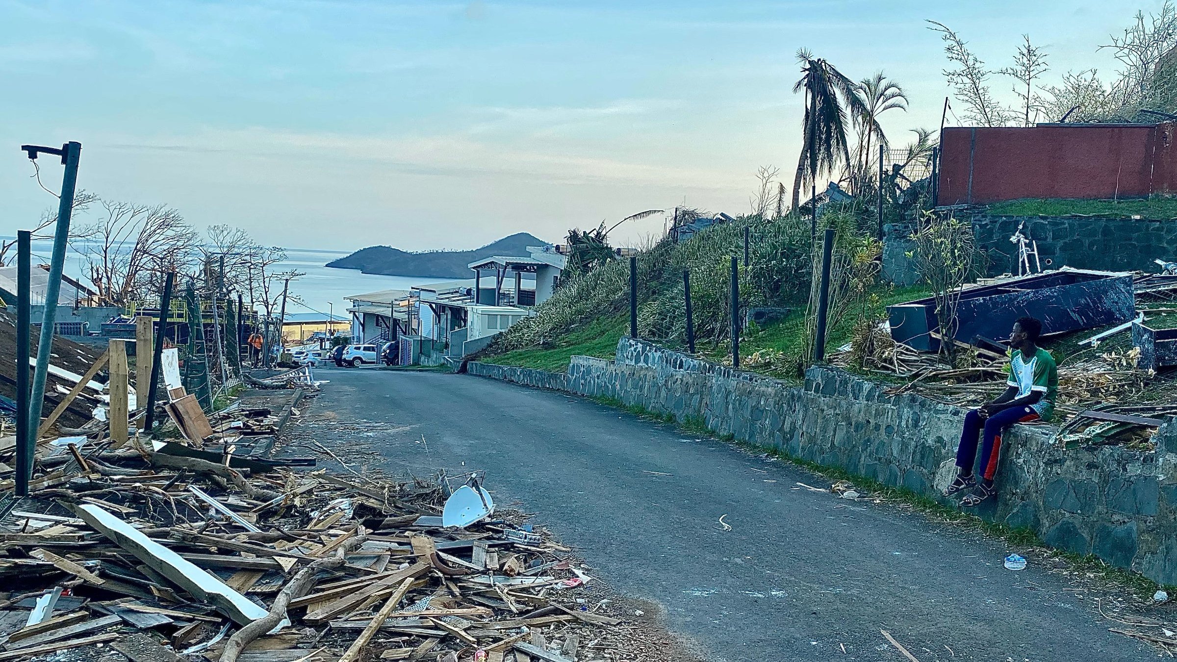 L'image présente une scène de destruction dans une rue. On voit des débris en bois éparpillés sur le sol, témoignant d'un événement dévastateur, peut-être une tempête ou un ouragan. Sur la gauche, il y a des maisons endommagées, certaines avec des toits manquants. En arrière-plan, on devine un paysage côtier avec une étendue d'eau qui reflète la lumière du ciel. À droite, une personne est assise sur un mur, habillée de vêtements colorés, semblant contempler les dégâts. La lumière du jour est douce, annonçant un crépuscule paisible, contrastant avec la morosité de la scène. Les palmiers, certains déracinés, ajoutent à l'impression de désolation.