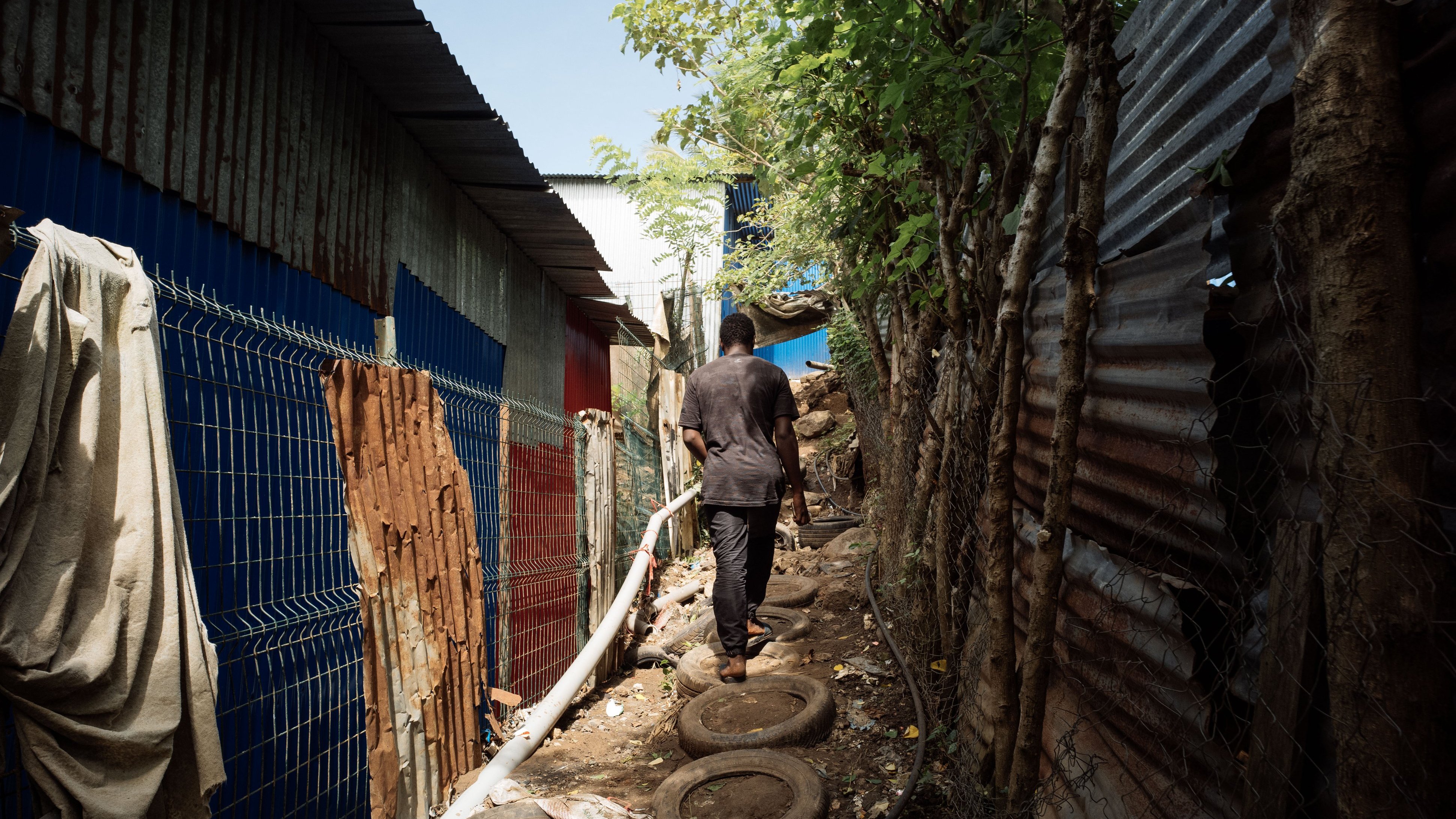 L'image montre un sentier étroit entre deux structures en tôle ondulée, typiques des bidonvilles. À gauche, on distingue des murs colorés, tandis qu'à droite, la structure est recouverte de tôles rouillées. Le chemin est constitué de pneus empilés, et un homme marche le long de ce chemin, portant des vêtements sombres. La lumière du jour illumine la scène, mettant en évidence la végétation qui borde le sentier, avec des arbres et des feuilles qui apportent une touche de verdure à cet environnement urbain. L'atmosphère semble à la fois animée et paisible.