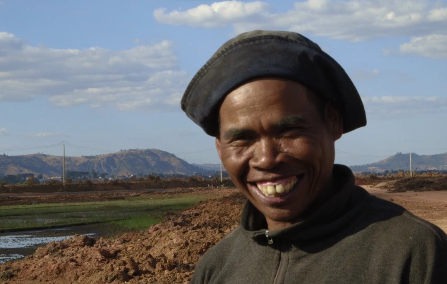 L'image montre un homme souriant se tenant devant un paysage rural. Il porte un chapeau noir et un vêtement sombre. En arrière-plan, on peut voir des collines et un ciel parsemé de nuages blancs. Le sol est terreux, et il y a des zones d'eau, probablement des rizières. L'expression de l'homme dégage de la joie et de la chaleur, ajoutant une touche positive à cette scène naturelle.