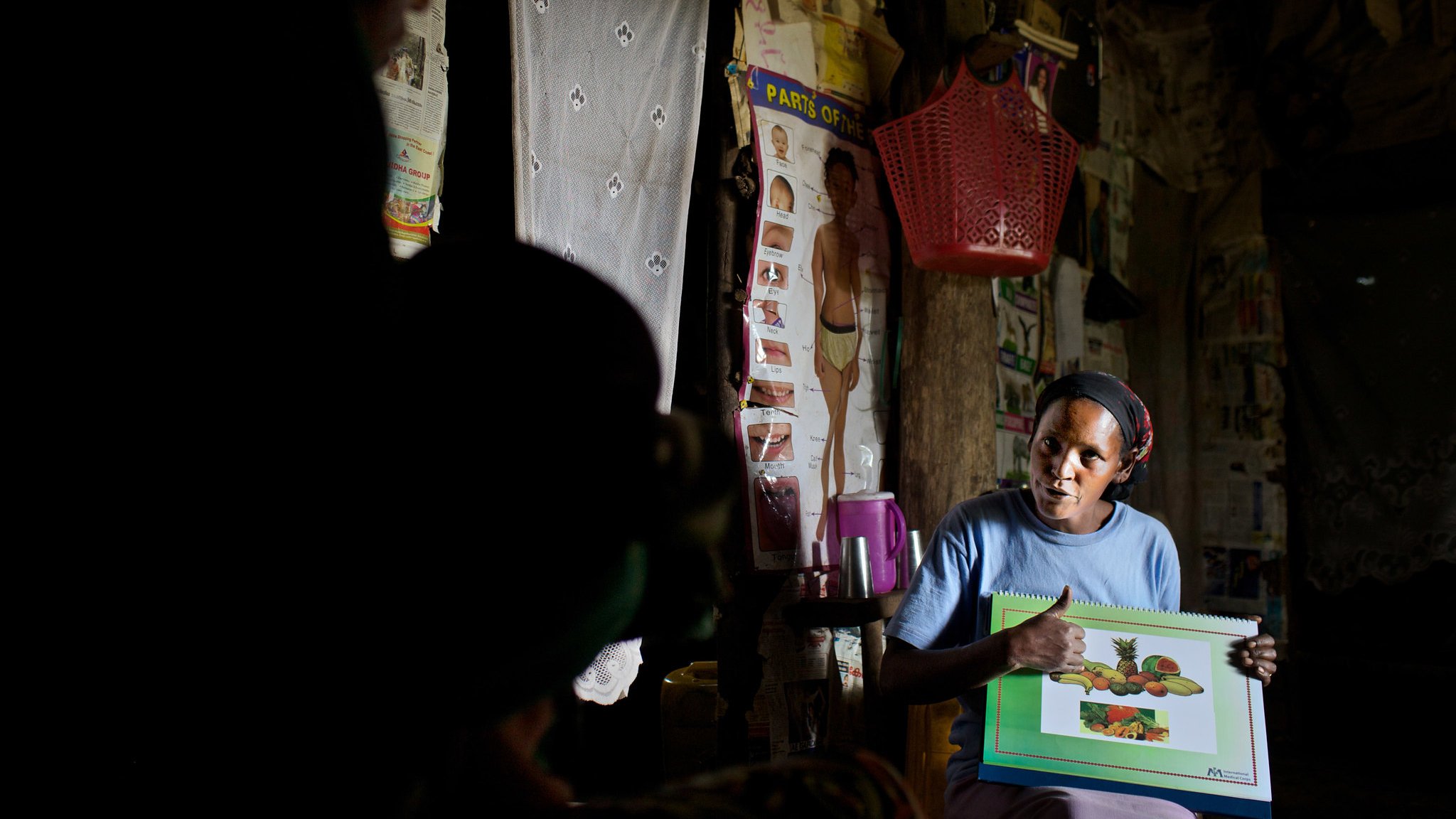 L'image montre une femme assise dans une pièce sombre, entourée de murs décorés d'affiches et de tissus. Elle tient un grand carnet avec des illustrations de fruits, qu'elle montre avec enthousiasme. Sa tenue est simple, et elle a l'air concentrée sur son explication. À l'arrière-plan, une silhouette d'une autre personne est visible, ajoutant à l'atmosphère d'échange. La scène évoque un moment d'apprentissage ou de partage d'informations sur les fruits.