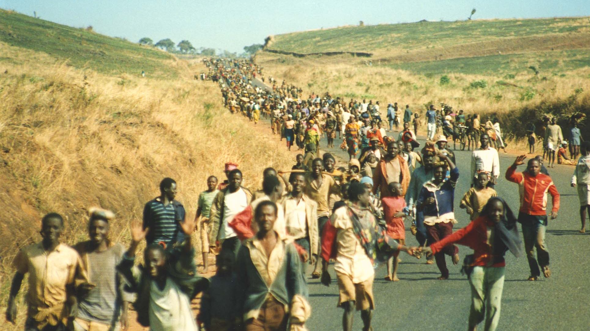 L'image montre une vaste foule de personnes marchant le long d'une route au milieu d'un paysage de collines sèches et herbeuses. Les individus, de différents âges et costumes, avancent ensemble, certains se tenant par la main ou portant des charges. L'atmosphère semble à la fois déterminée et solennelle, peut-être symbolisant un déplacement ou une migration collective. En arrière-plan, on aperçoit une étendue de terre vallonnée qui s'étend à perte de vue, ajoutant une dimension de profondeur à la scène. Le ciel est dégagé, laissant entrevoir un jour ensoleillé.