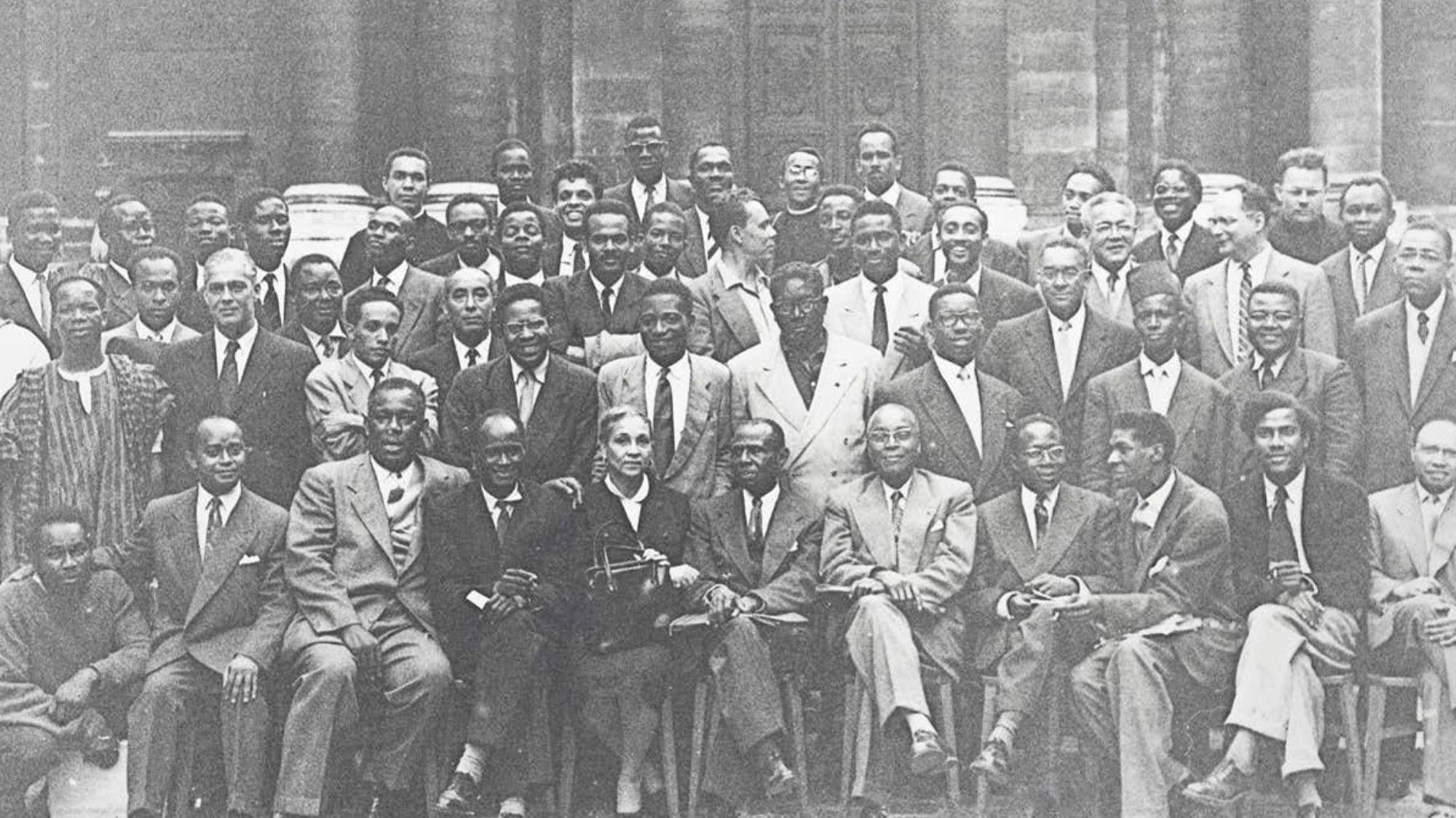 L'image présente un groupe de plusieurs hommes, probablement des leaders et des intellectuels, rassemblés pour une photographie de groupe. Ils sont vêtus de costumes formels, avec une variété de poses : certains sont assis, d'autres debout. L'ambiance semble chaleureuse et conviviale. Au centre, une femme se distingue, assise avec un sourire. En arrière-plan, on aperçoit un bâtiment historique, suggérant peut-être un événement ou une rencontre importante. L'image est en noir et blanc, ce qui lui donne un aspect intemporel.