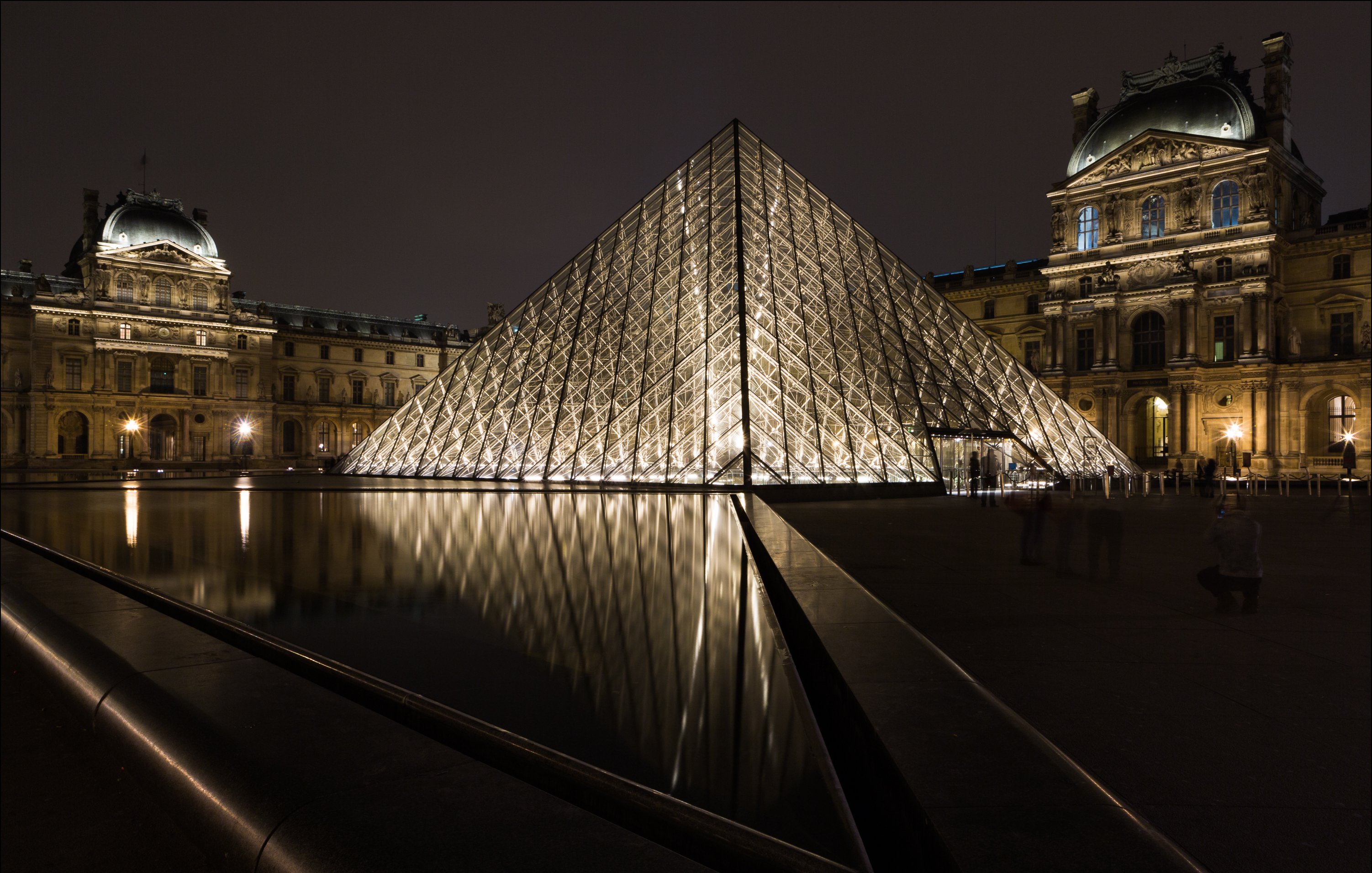 L'image montre le célèbre musée du Louvre à Paris, avec sa pyramide en verre illuminée au centre de la scène. La pyramide, qui est moderne et de forme géométrique, se dresse majestueusement au-dessus d'un bassin d'eau calme qui en reflète la lumière. À gauche, on aperçoit l'architecture historique du musée, avec ses pierres anciennes et des détails raffinés. La scène est plongée dans une ambiance nocturne, où les lumières chatoyantes se mêlent à l'obscurité, créant un contraste saisissant entre le moderne et le classique. Le reflet dans l'eau ajoute une dimension poétique à l'ensemble.