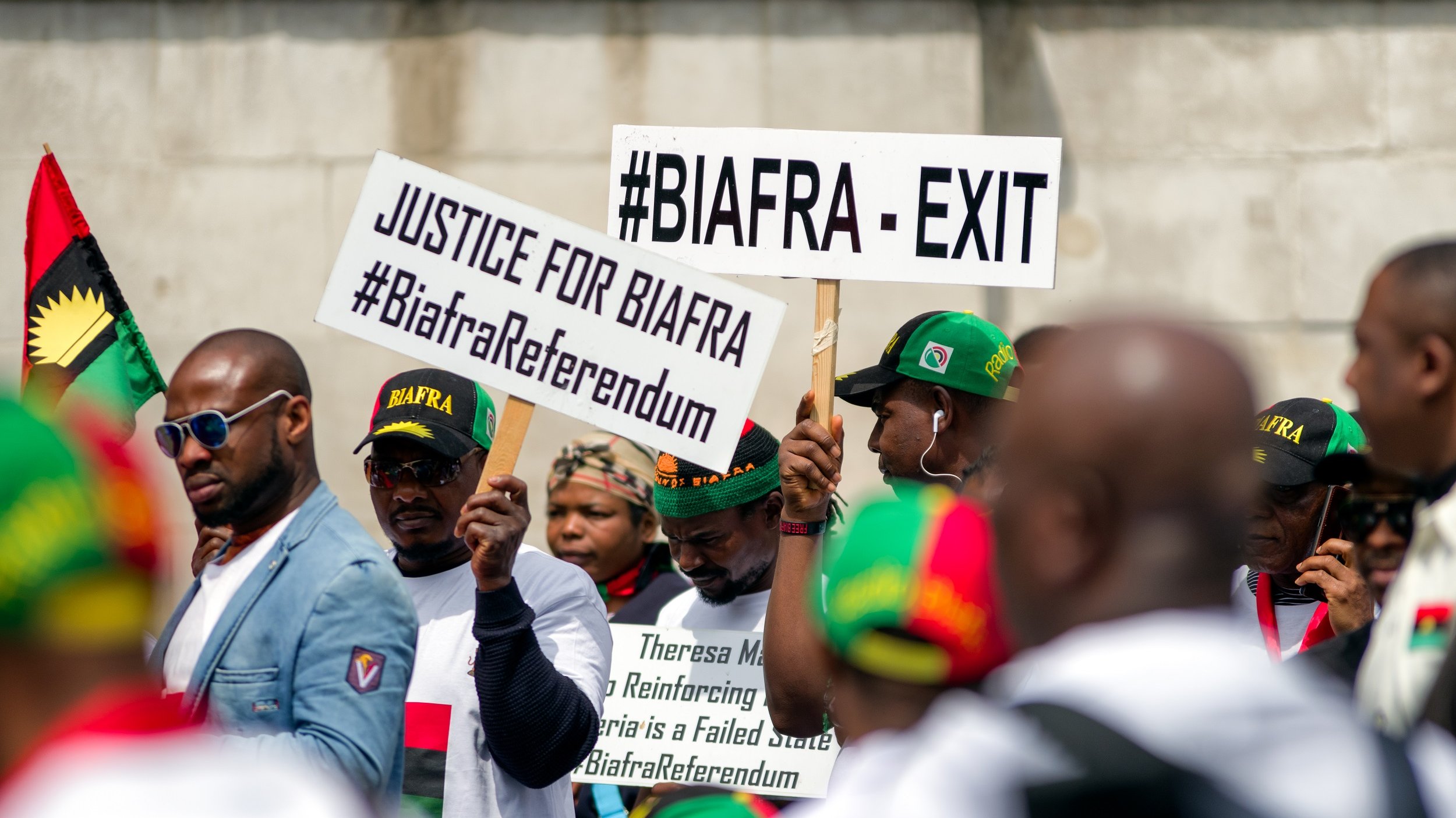 L'image montre un groupe de personnes participant à une manifestation. Elles portent des t-shirts blancs et des casquettes aux couleurs du mouvement Biafra. Des pancartes sont brandies, portant des inscriptions telles que "#BIAFRA - EXIT" et "JUSTICE FOR BIAFRA", ainsi que des hashtags liés au référendum biafrais. L'atmosphère semble dynamique et engagée, reflétant une revendication pour la justice et l'autodétermination.