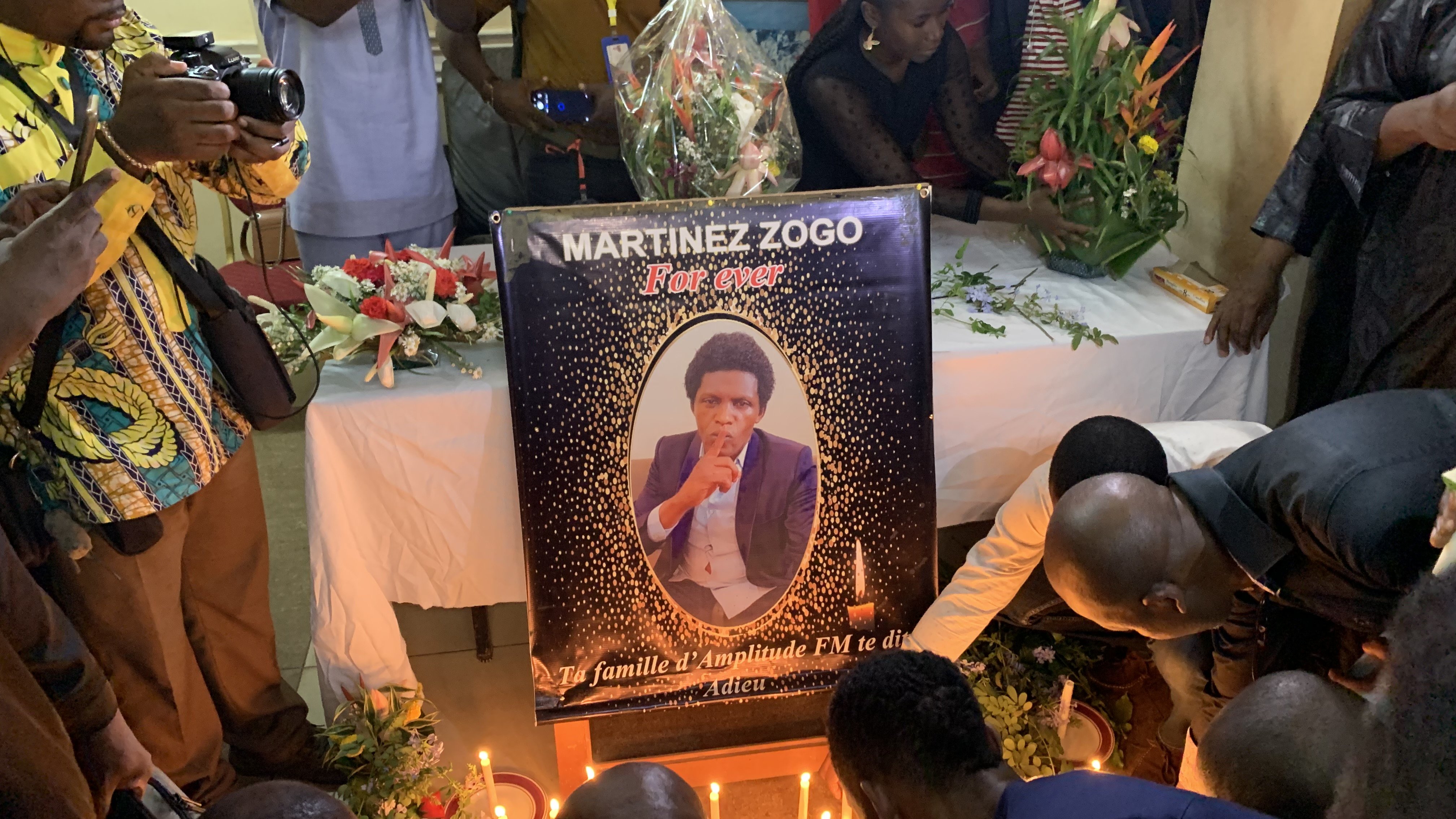 L'image dépeint une scène de recueillement. Au centre, il y a un grand poster représentant Martinez Zogo, encadré de fleurs et de bougies allumées. Le poster montre une photo de lui en train de fumer une cigarette, avec un regard pensif. Autour du poster, plusieurs personnes sont rassemblées, certaines s'agenouillant ou se penchant pour prier. L'atmosphère est solennelle, avec des expressions de tristesse sur les visages. Des fleurs et des bougies décorent la table sur laquelle le poster repose, ajoutant une touche de commémoration à cet hommage.