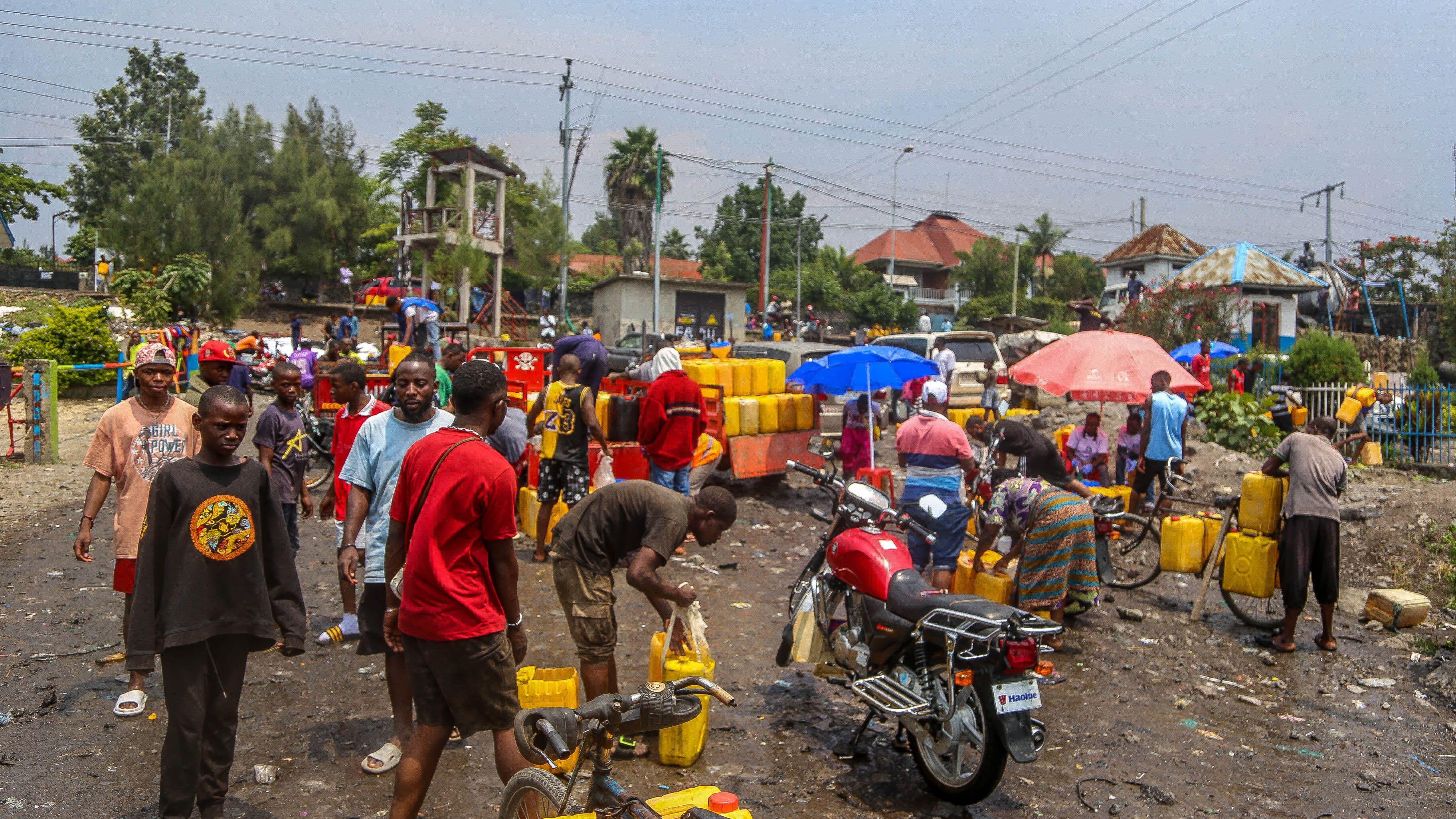 L'image représente une scène animée dans un quartier urbain. On peut imaginer un marché ou un point de distribution d'eau, où de nombreuses personnes sont rassemblées. Des hommes et des femmes, de divers âges, s'affairent autour de grands récipients jaunes qu'ils remplissent d'eau. Le sol semble en désordre, avec des déchets visibles, ajoutant une impression de négligence dans l'environnement. Des parapluies colorés sont déployés, fournissant un peu d'ombre sous un ciel légèrement nuageux. Au fond, on peut apercevoir des bâtiments et des arbres, tandis qu'un motard fait la queue avec sa moto, indiquant une activité routière environnante. L'atmosphère est dynamique, avec une forte interaction entre les gens.