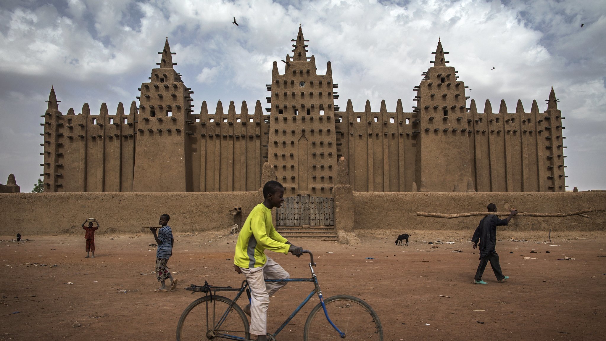 L'image montre une grande mosquée en banco, typique de l'architecture soudanaise, avec des murs en terre crue qui s'élèvent vers le ciel. Sa façade est ornée de nombreuses pointes et créneaux, formant un motif impressionnant. Au premier plan, un jeune garçon traverse la scène à bicyclette, tandis que d'autres enfants jouent et se déplacent dans l'espace. Le sol est poussiéreux, et le climat semble chaud, avec des nuages ici et là dans le ciel. L'ambiance est vivante, illustrant une scène animée de la vie quotidienne.
