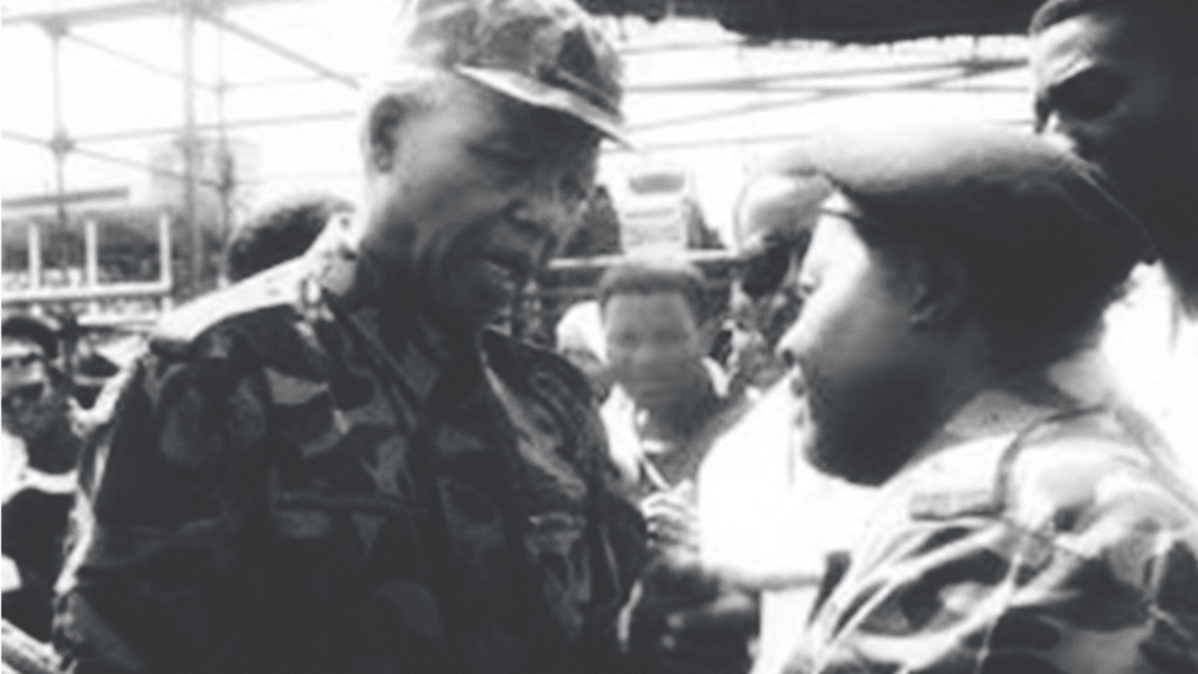 Cette image en noir et blanc montre deux hommes engagés dans une conversation intense. L'homme à gauche, avec un chapeau de camouflage et un uniforme assorti, a une expression sérieuse et attentive. À sa droite, un autre homme portant également une tenue de camouflage semble écouter avec intérêt. En arrière-plan, on peut voir un groupe de personnes, floues, qui regardent la scène. L'atmosphère de l'image suggère un moment significatif, probablement lié à une discussion importante ou à un événement marquant.