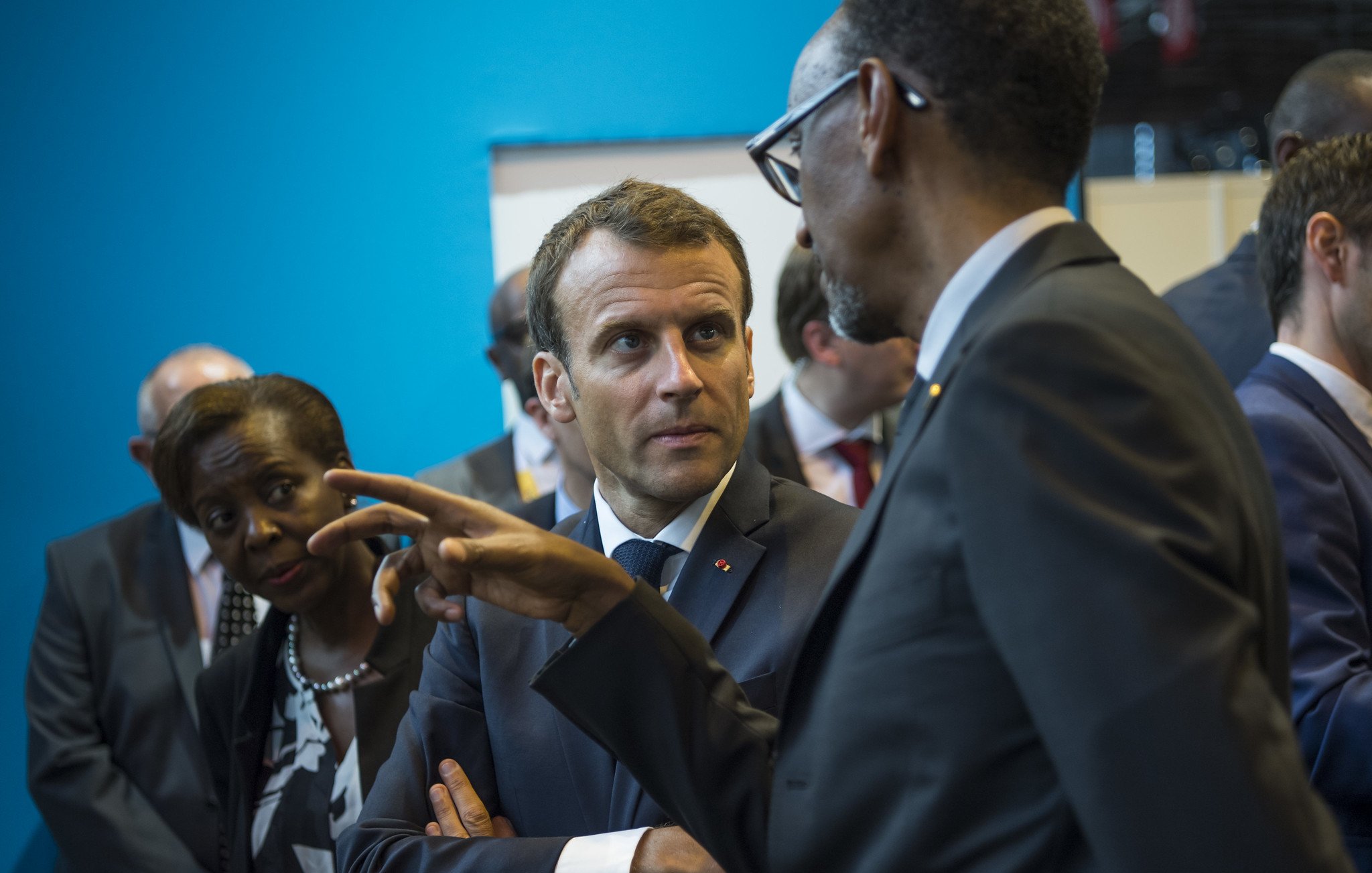 L'image montre un groupe de personnes en conversation dans un environnement qui semble formel. Au premier plan, un homme en costume sombre, probablement un leader, discute avec un autre homme, qui porte également un manteau formel. Le premier homme a une expression attentive, se concentrant sur la discussion, tandis que le second gesticule avec sa main, comme s'il faisait un point important. En arrière-plan, une femme les observe, affichant une expression de sérieux. Le décor est composé de murs d'un bleu vif, ce qui crée un contraste avec les costumes sombres des personnages. L'atmosphère semble professionnelle et engagée, suggérant une rencontre importante.