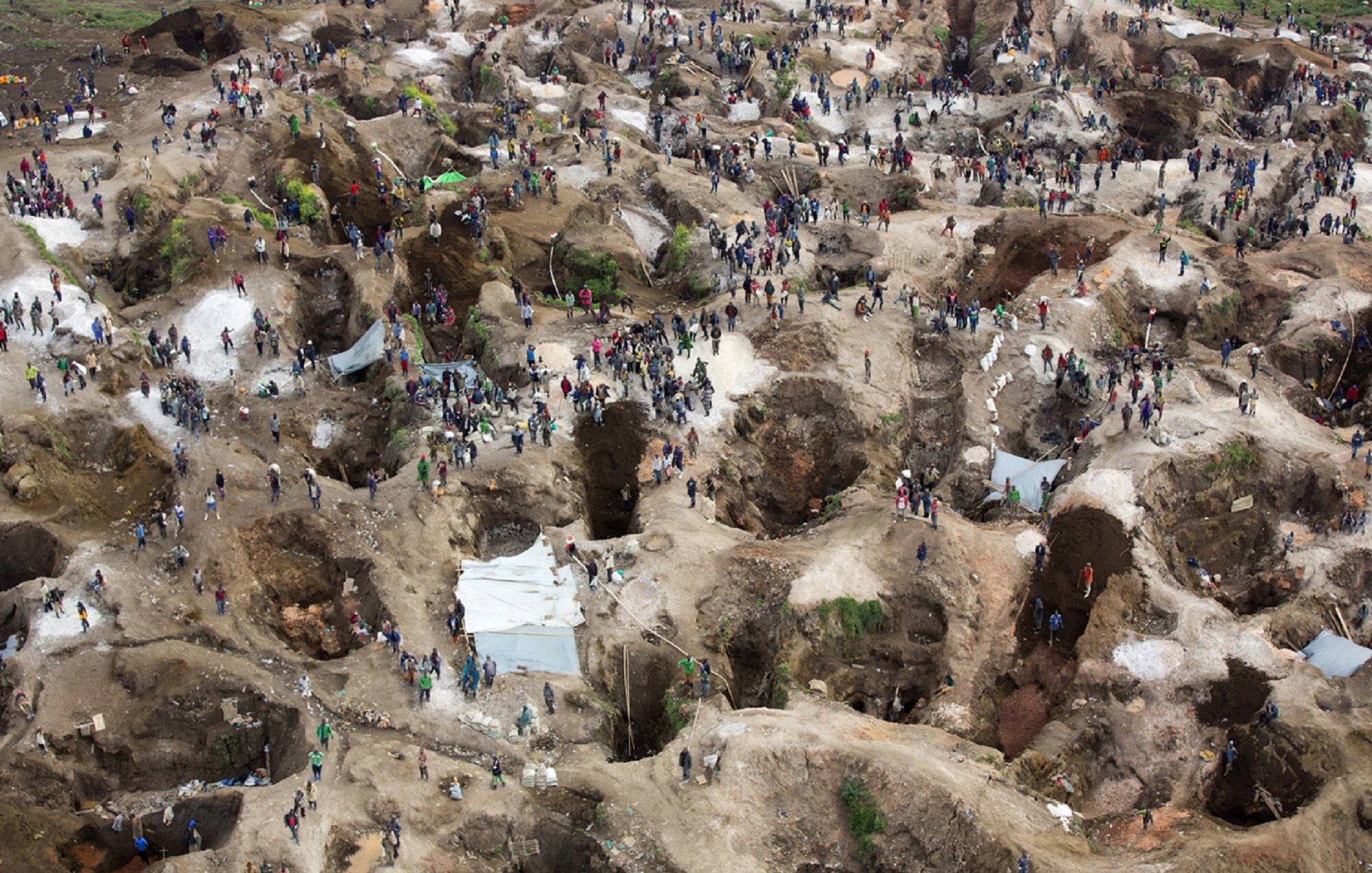 L'image représente une grande site d'extraction minière, probablement un lieu de prospection. On y voit de nombreuses personnes, qui semblent être des mineurs ou des chercheurs de minerai, dispersées sur un terrain accidenté. Le sol est parsemé de nombreuses cavités, témoignant des efforts de creusement. Certaines zones sont abritées par des abris temporaires en toile, tandis que d'autres sont à ciel ouvert. La scène dégage une dynamique de travail acharné, où les individus s'affairent dans un environnement à la fois désordonné et organisé, caractérisé par des monticules de terre et des roches. L'ensemble évoque un lieu de quête et de lutte pour des ressources précieuses.