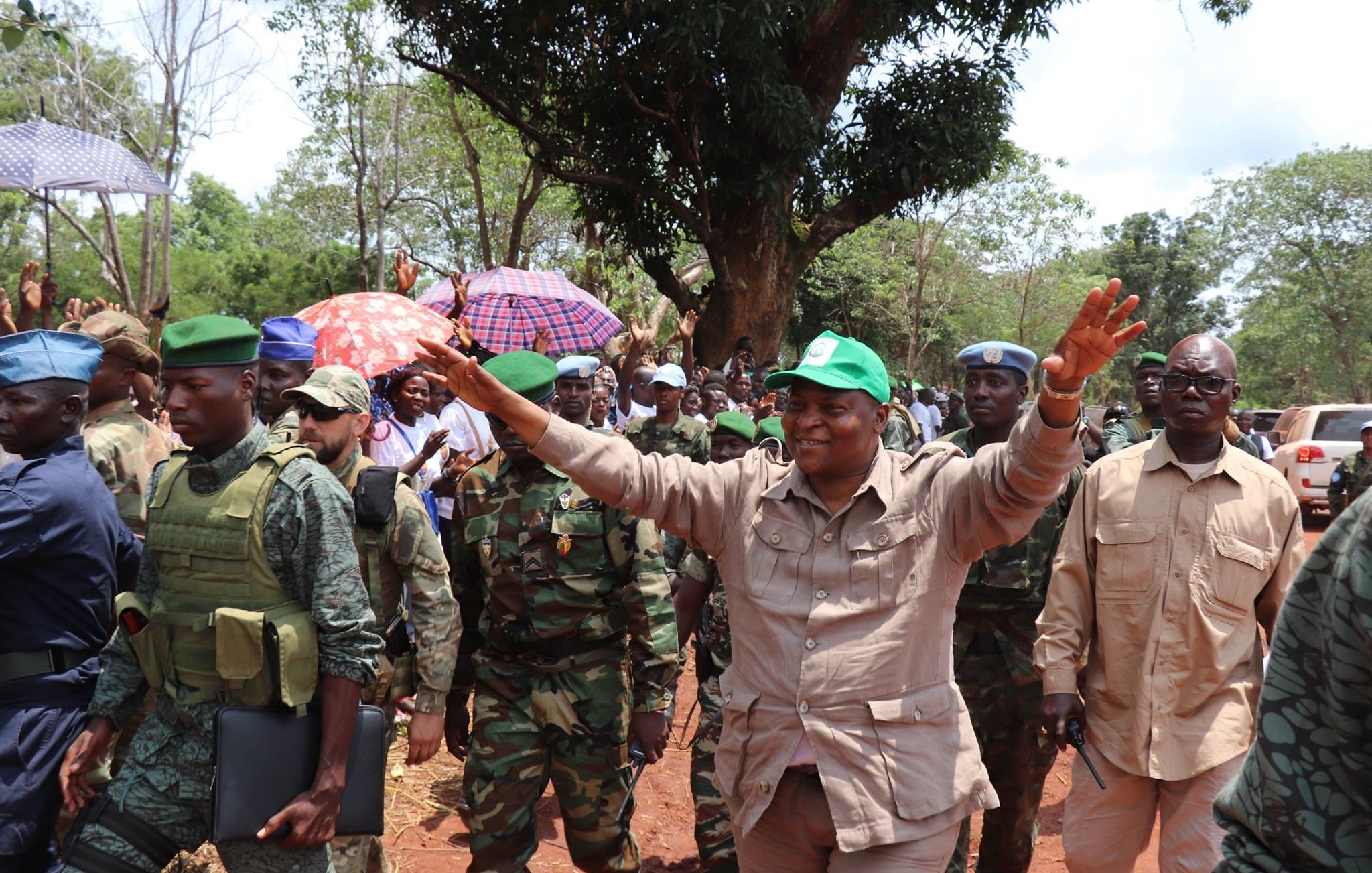 L'image montre une scène animée en plein air, avec un groupe de personnes réunies. Au centre, un homme souriant, vêtu d'une tenue beige et portant une casquette verte, semble saluer la foule avec les bras levés. Autour de lui, plusieurs militaires en uniforme, certains portant des gilets pare-balles, se tiennent en rang. La foule en arrière-plan est composée de personnes sous des parapluies, suggérant une atmosphère festive ou de célébration. Des arbres verdoyants et un chemin de terre sont visibles, créant un cadre naturel et vivant. L'ambiance semble énergique et encourageante.