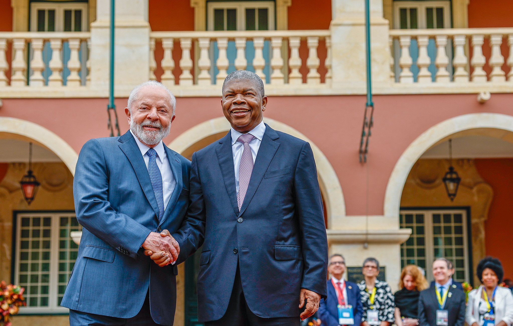 L'image montre deux hommes se serrant la main, symbolisant un moment de convivialité et de coopération. En arrière-plan, on peut apercevoir une architecture colorée avec des colonnes et des balcons. Des personnes sont rassemblées à l'arrière, témoignant d'un événement officiel ou d'une rencontre importante. Les vêtements des hommes sont élégants : l'un porte un costume gris et l'autre un costume noir avec une cravate claire. L'atmosphère semble chaleureuse et dynamique, reflétant un échange amical.
