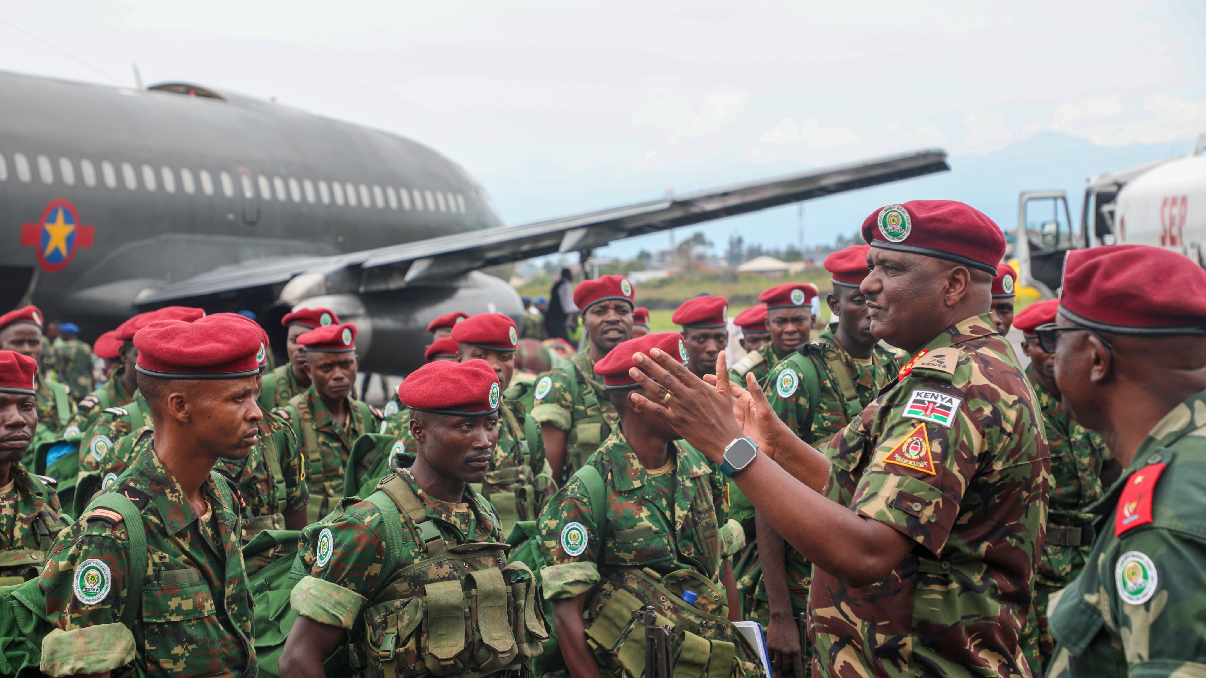 L'image montre un groupe de soldats en uniforme militaire, réunis près d'un gros avion de transport. Les soldats portent des casques et des berets rouges, et ils sont habillés de camouflages verts. L'un des officiers, qui semble donner des instructions ou des encouragements, se tient au centre, gesticulant avec les mains. En arrière-plan, on peut voir l'avion de taille importante, avec des couleurs de camouflage, sur un terrain d'aviation, tandis que le ciel est légèrement nuageux. L'atmosphère est sérieuse, témoignant d'une mobilisation militaire.
