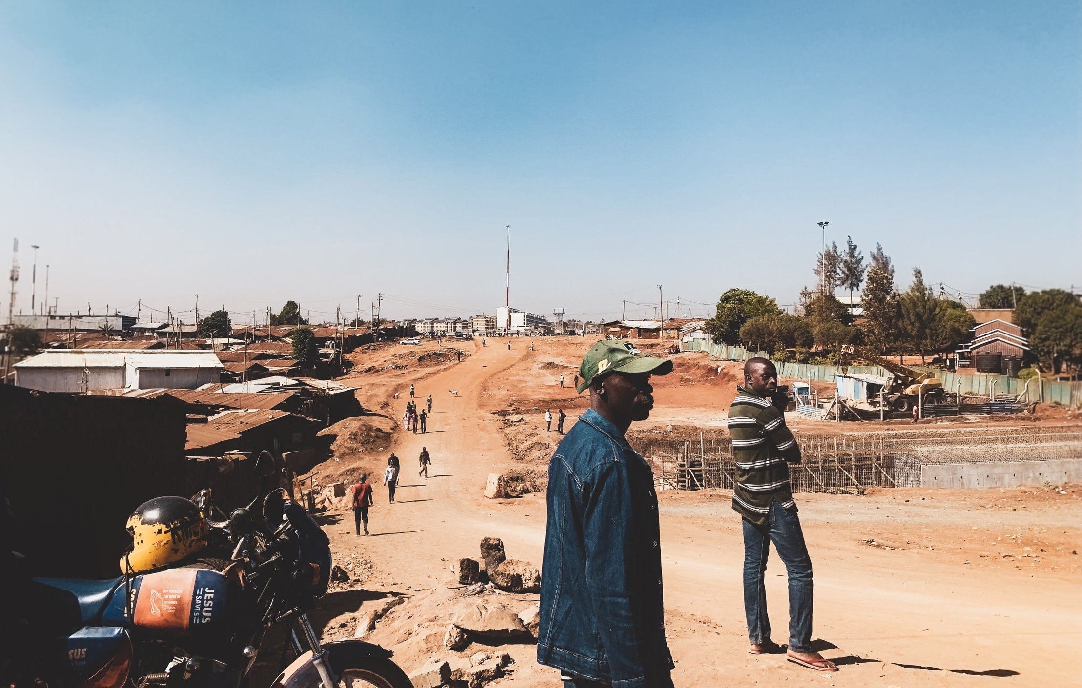 Dans cette image, nous voyons une scène d'une rue en construction dans une zone urbaine. Le ciel est dégagé et d'un bleu clair. Au premier plan, deux hommes se tiennent debout : l'un porte une veste en jean et une casquette, tandis que l'autre est vêtu d'un sweater rayé. À droite, on aperçoit un chemin de terre, entouré de bâtiments en dur et d'habitations simples. À gauche, une moto est garée, et plusieurs personnes se déplacent le long de la route. L'atmosphère semble calme, mais l'environnement indique une activité en cours avec des travaux de construction en arrière-plan.