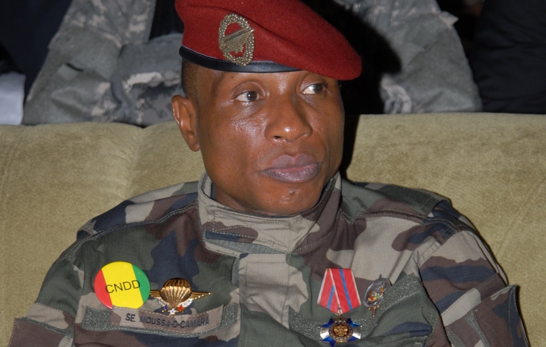 L'image montre un militaire assis, vêtu d'un uniforme camouflage. Il porte une casquette rouge ornée d'un insigne. Sur sa poitrine, il y a plusieurs décorations et médailles visibles. Les couleurs de l'uniforme et des insignes, notamment le jaune et le rouge, évoquent des éléments de son pays. Son expression est sérieuse, et il semble attentif, probablement à une discussion ou à un événement. L'arrière-plan est flou, suggérant une atmosphère formelle ou solennelle.