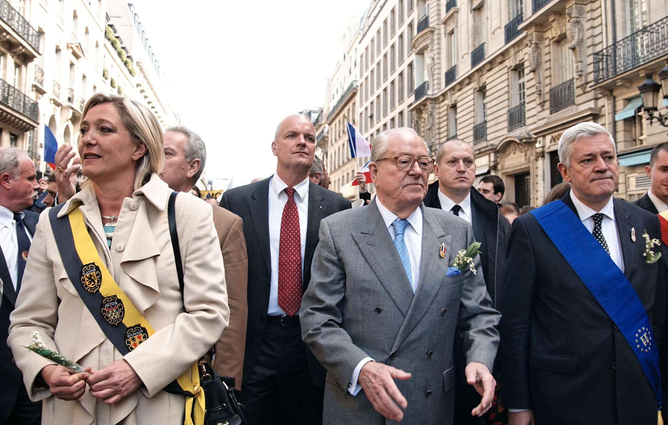 L'image montre un groupe de personnes marchant dans une rue animée, probablement lors d'un événement ou d'une cérémonie. Au centre, un homme plus âgé porte un costume gris clair et des lunettes. Il semble être une figure de proue, entouré de plusieurs autres individus, dont certains portent des sashes ou des écharpes distinctives, suggérant qu'ils occupent des rôles importants. On remarque également des décorations florales, indiquant une atmosphère cérémonielle. En arrière-plan, on aperçoit des bâtiments typiques, et des drapeaux flottent, ce qui évoque un rassemblement public festif et patriotique.
