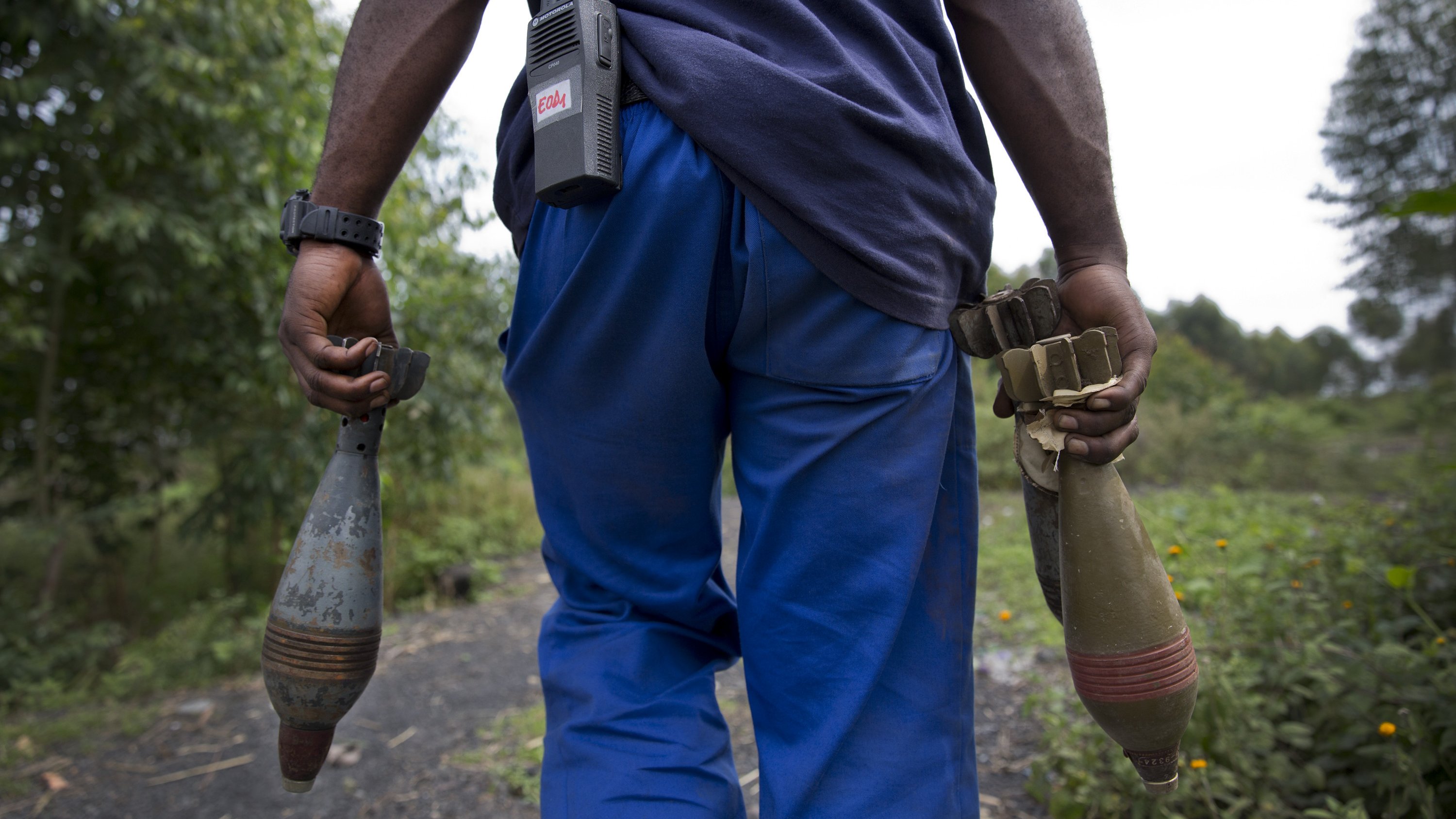 L'image montre un homme vu de dos, portant des vêtements décontractés : un t-shirt bleu foncé et un pantalon bleu. Il tient dans chaque main un objet long et cylindrique, probablement des munitions ou des dispositifs explosifs. À sa taille, il y a un appareil de communication. En arrière-plan, on aperçoit une nature verdoyante, avec des arbres et des plantes. L'ambiance semble à la fois sérieuse et chargée d'une importance qui nécessite prudence et vigilance.